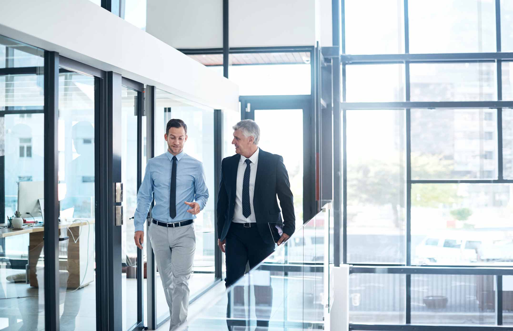 Two businessmen discussing resilience while walking in a modern office hallway with large windows.
