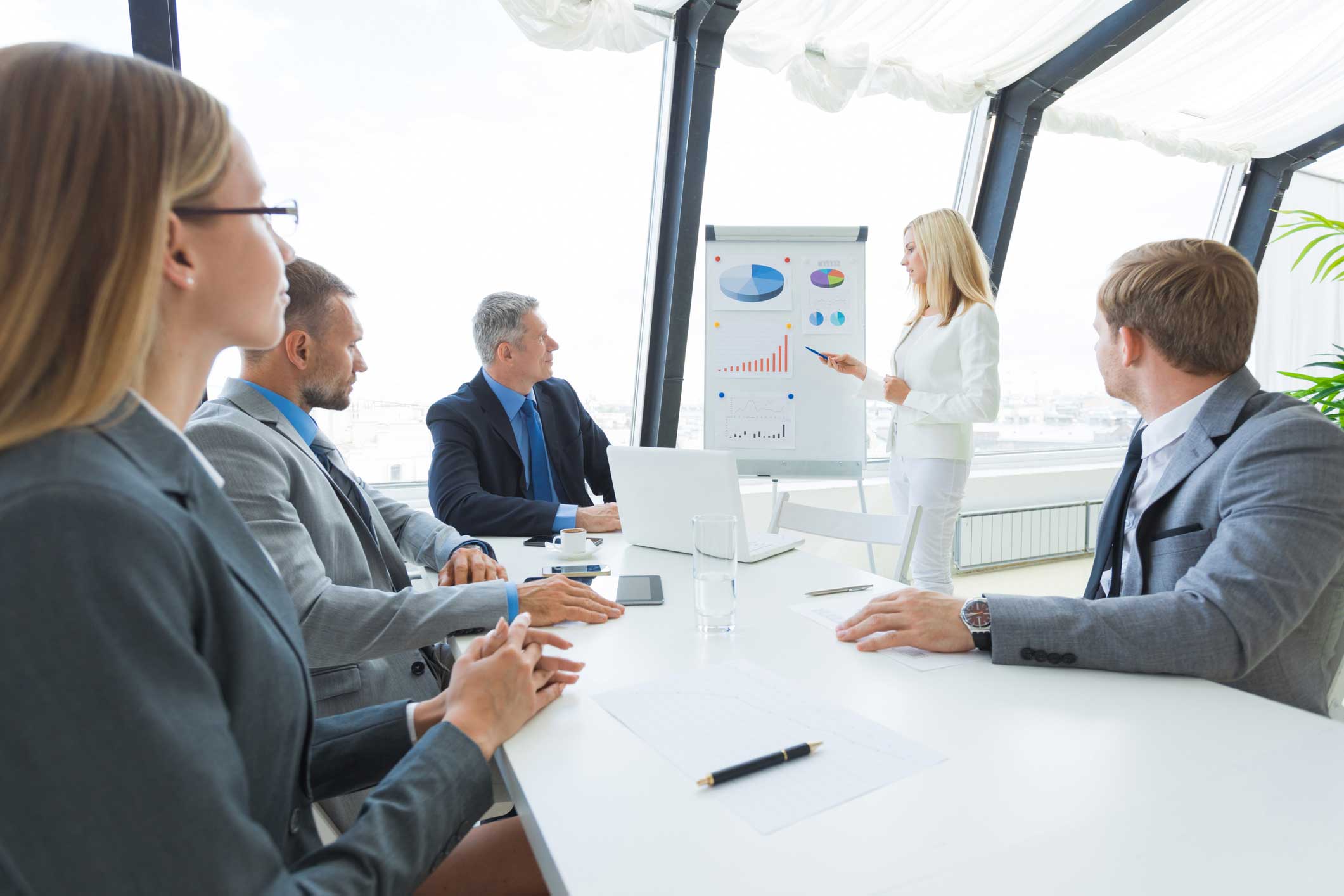 Four businesspeople hear a woman present charts on profitability intelligence in a modern office.