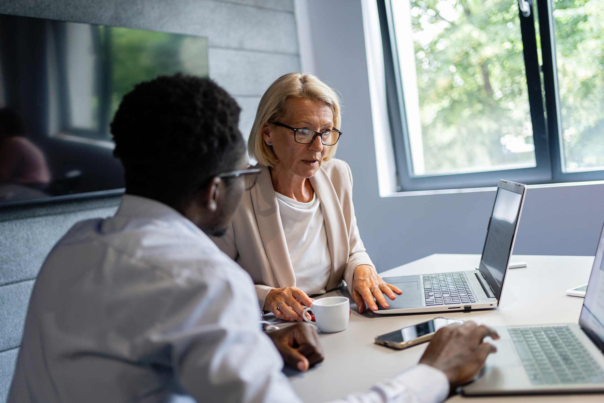 Two information security consultants discuss at a table with laptops, phone, and coffee cup.