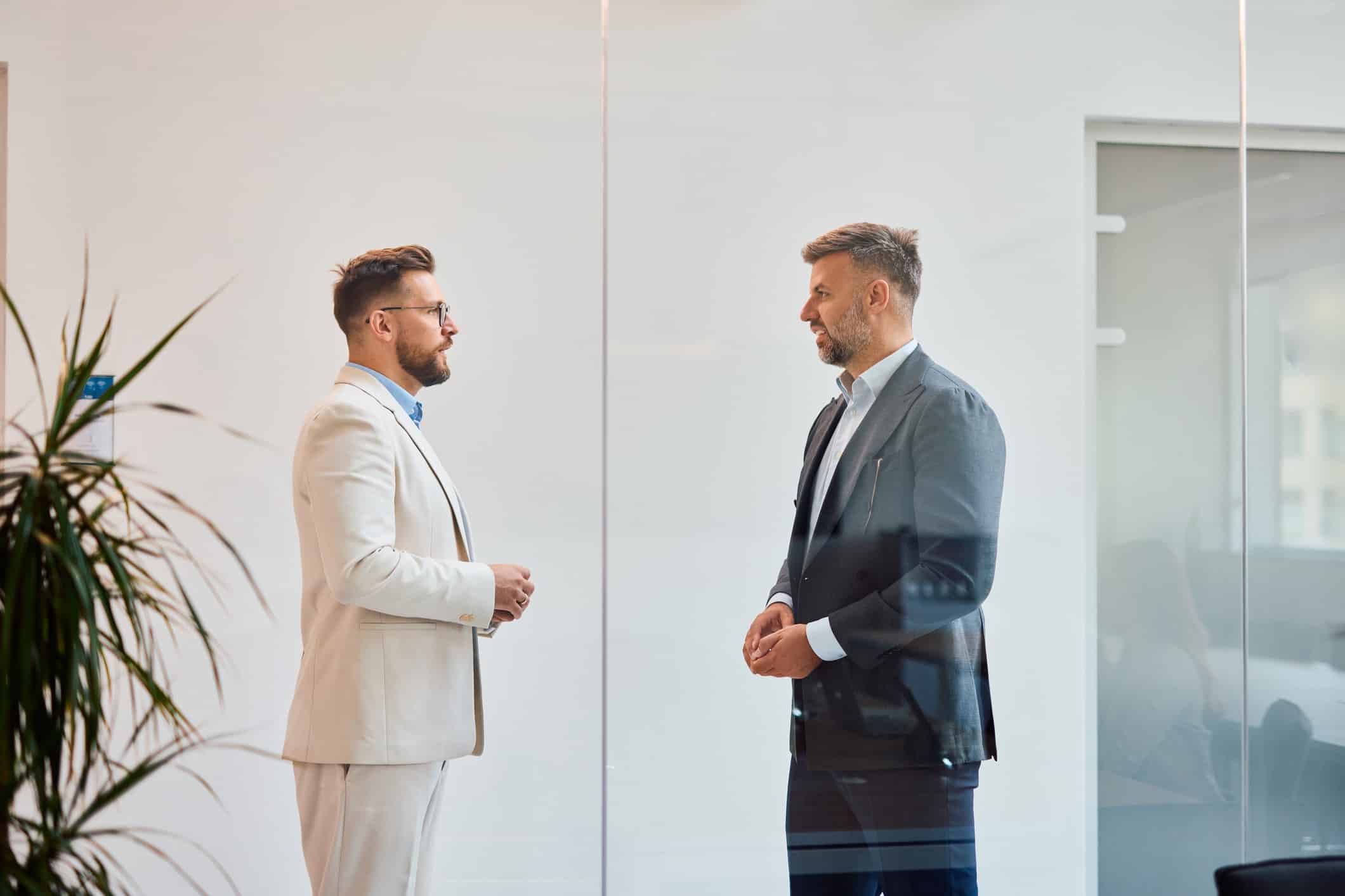 Two men in suits discuss in a modern office at a competitive strategy consulting firm.