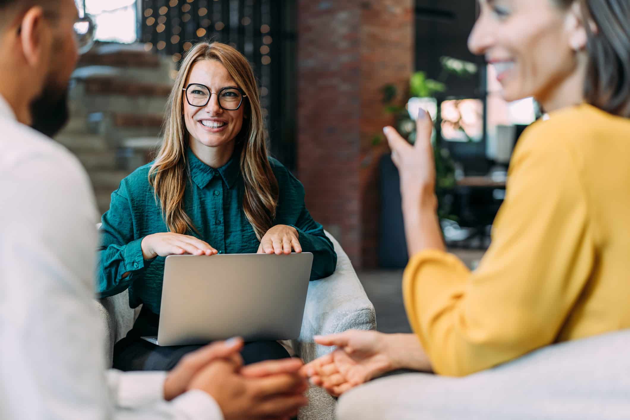 Three people talk in an office; one woman smiles with a laptop at a law firm advisory meeting.