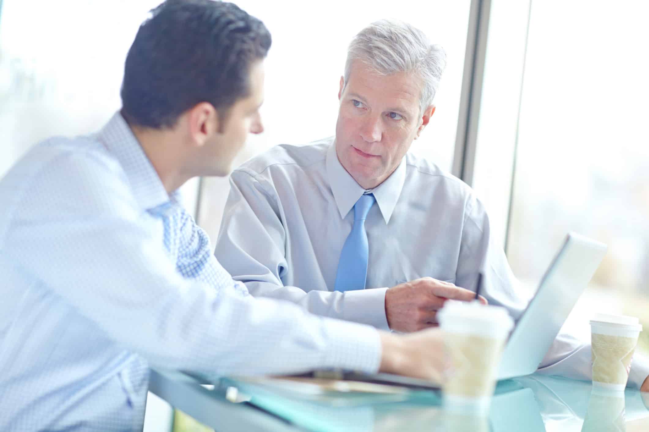 Two men in business attire discuss Law Firm Financial Advisory at a table with laptop and coffee.
