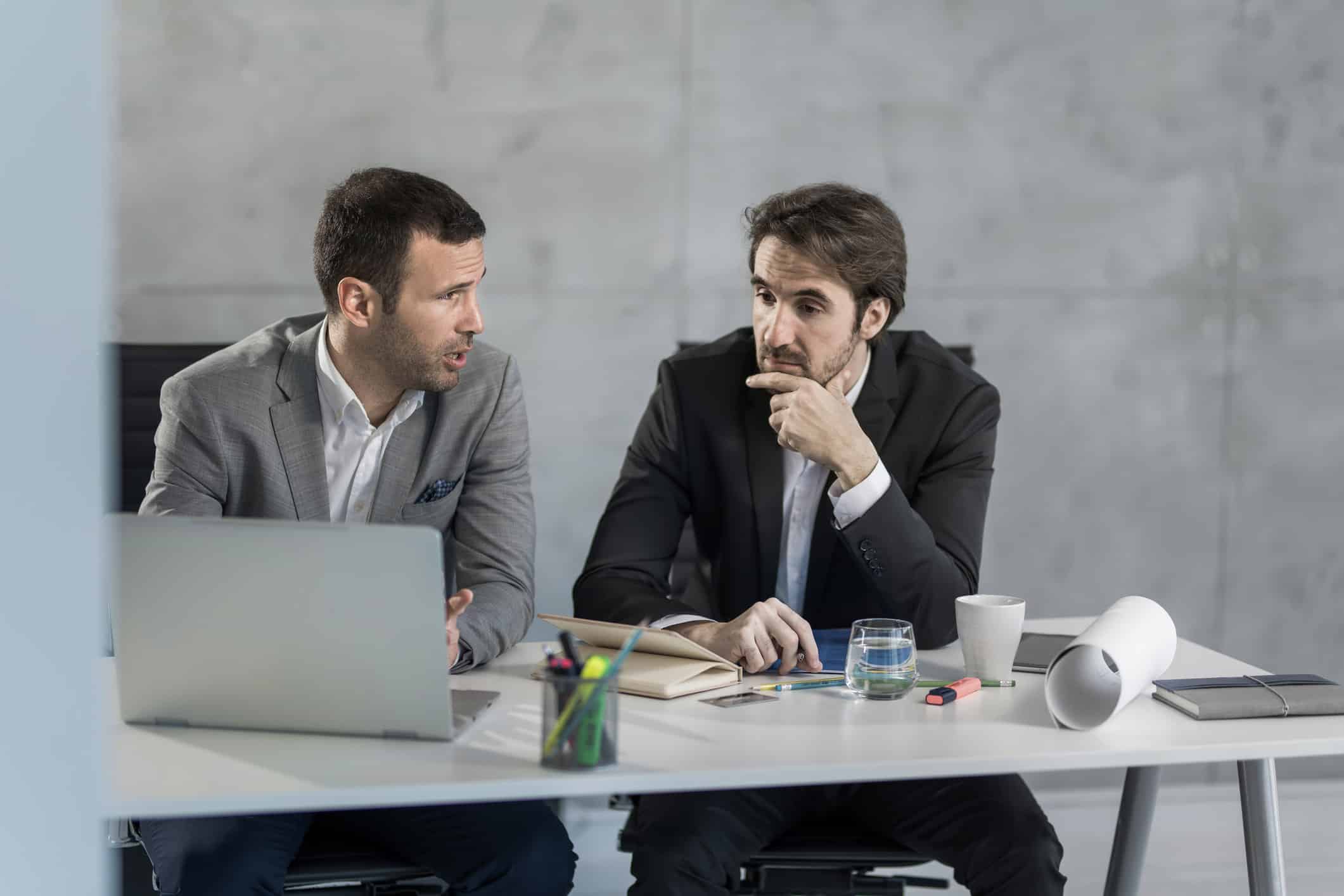 Two men in suits discuss corporate tax consulting at a desk with a laptop and office supplies.