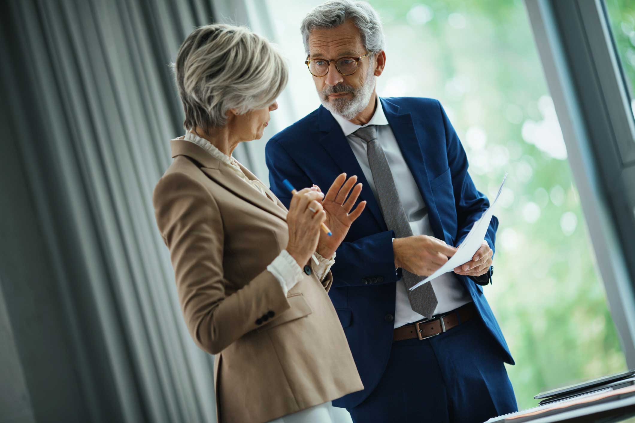 Two business professionals discuss it disaster recovery advisory by a window, holding documents.