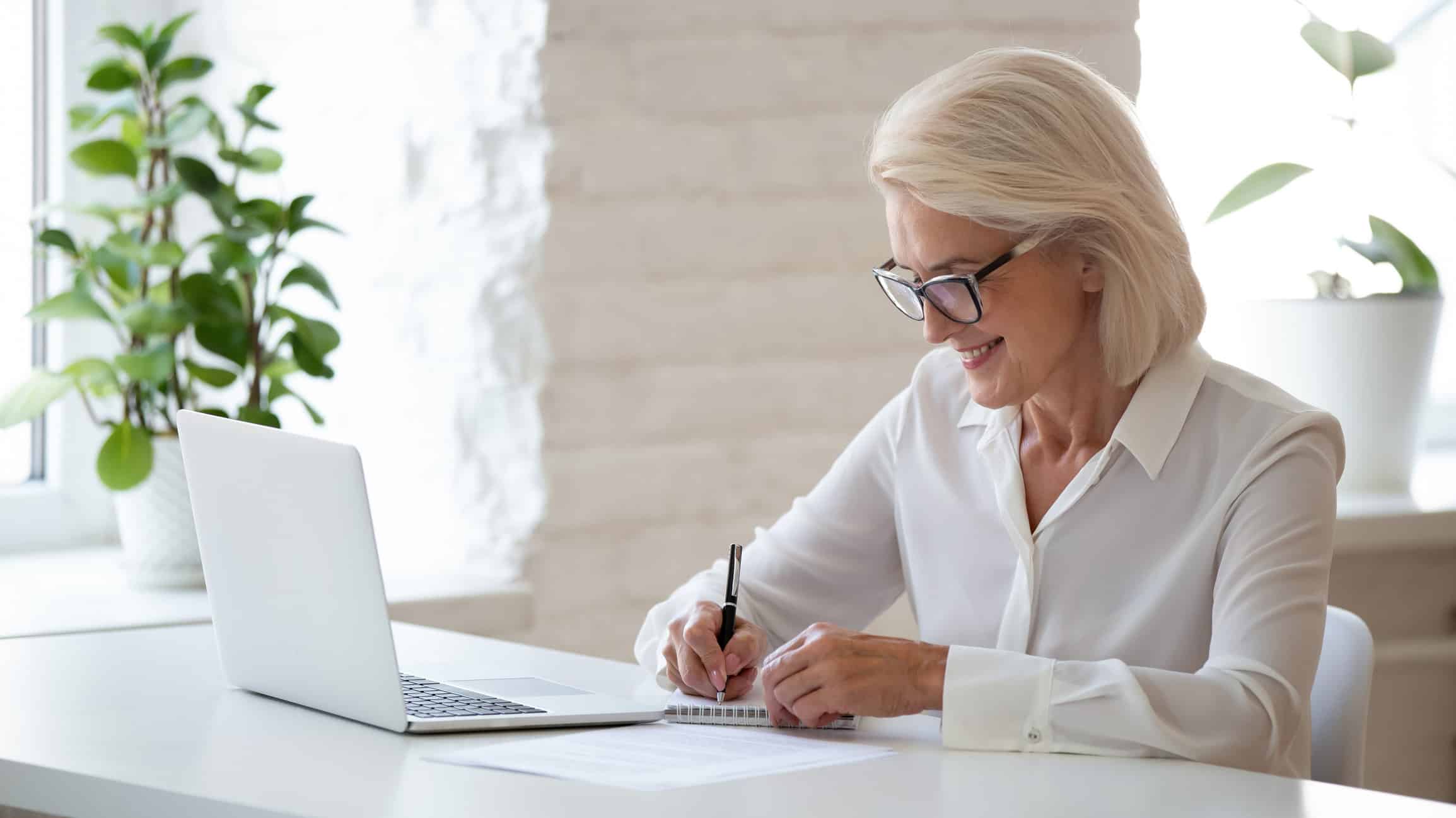 Smiling older woman in glasses writing about Antitrust Economic issues at a desk with a laptop.