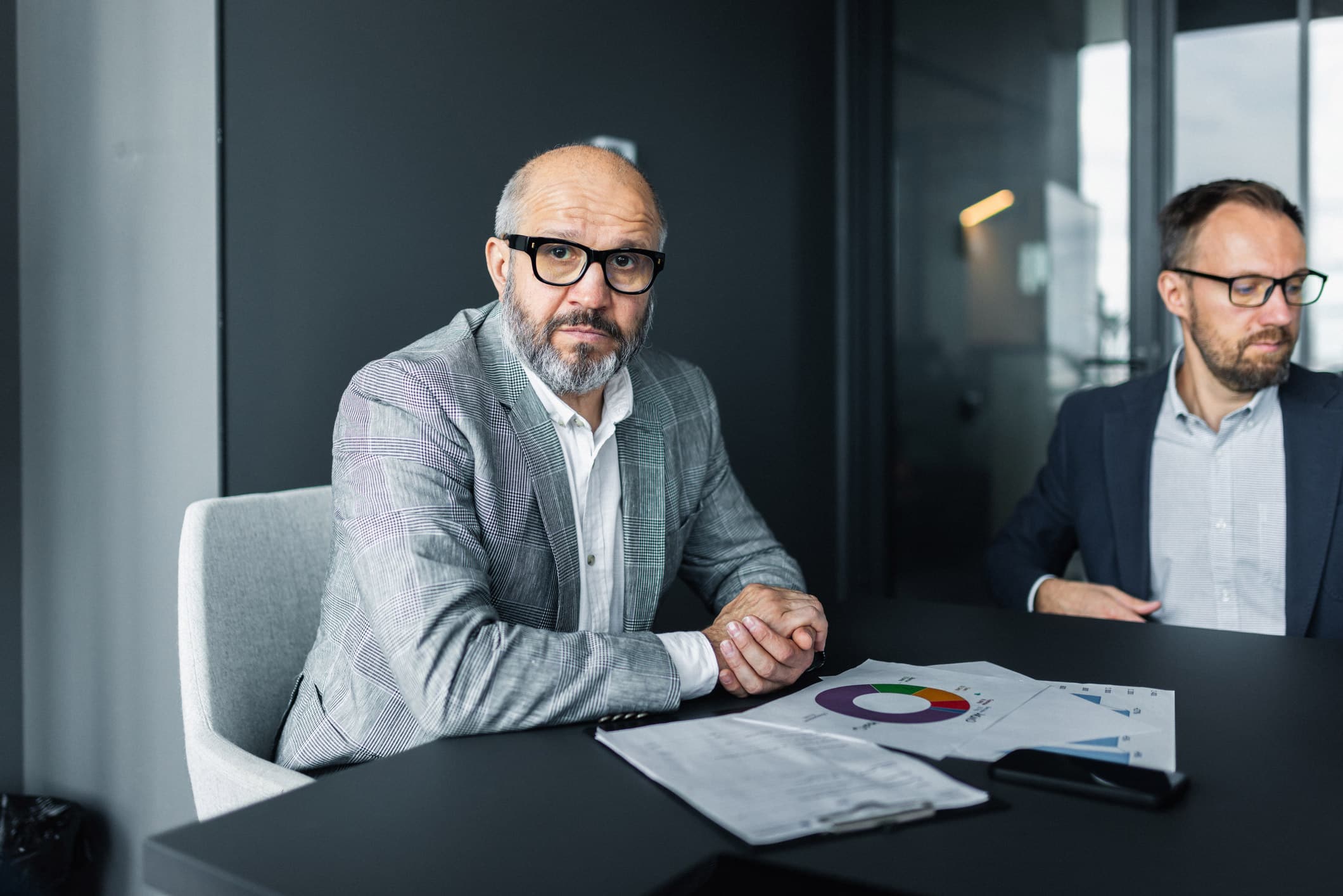 Two men in business attire discuss Risk Management Advisory at table with charts in an office.