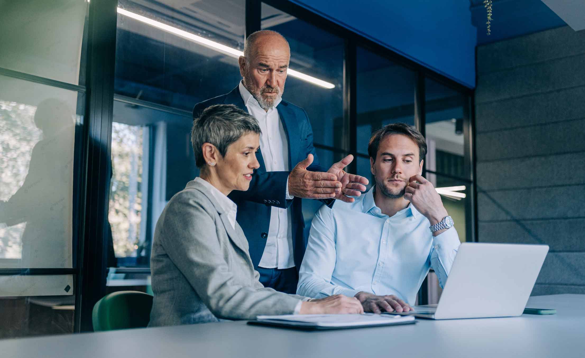 Three colleagues discuss AI Legal Document Analysis Consulting Services by a laptop in an office.