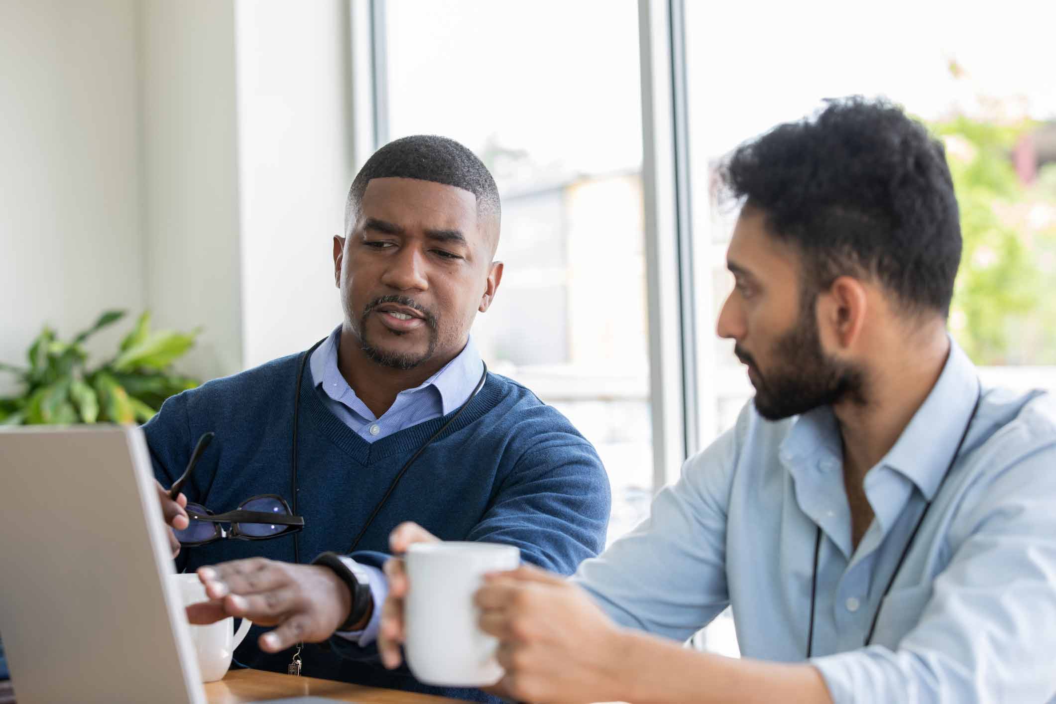 Two men sit at a table, coffee in hand, as data breach consultants discuss something on a laptop.