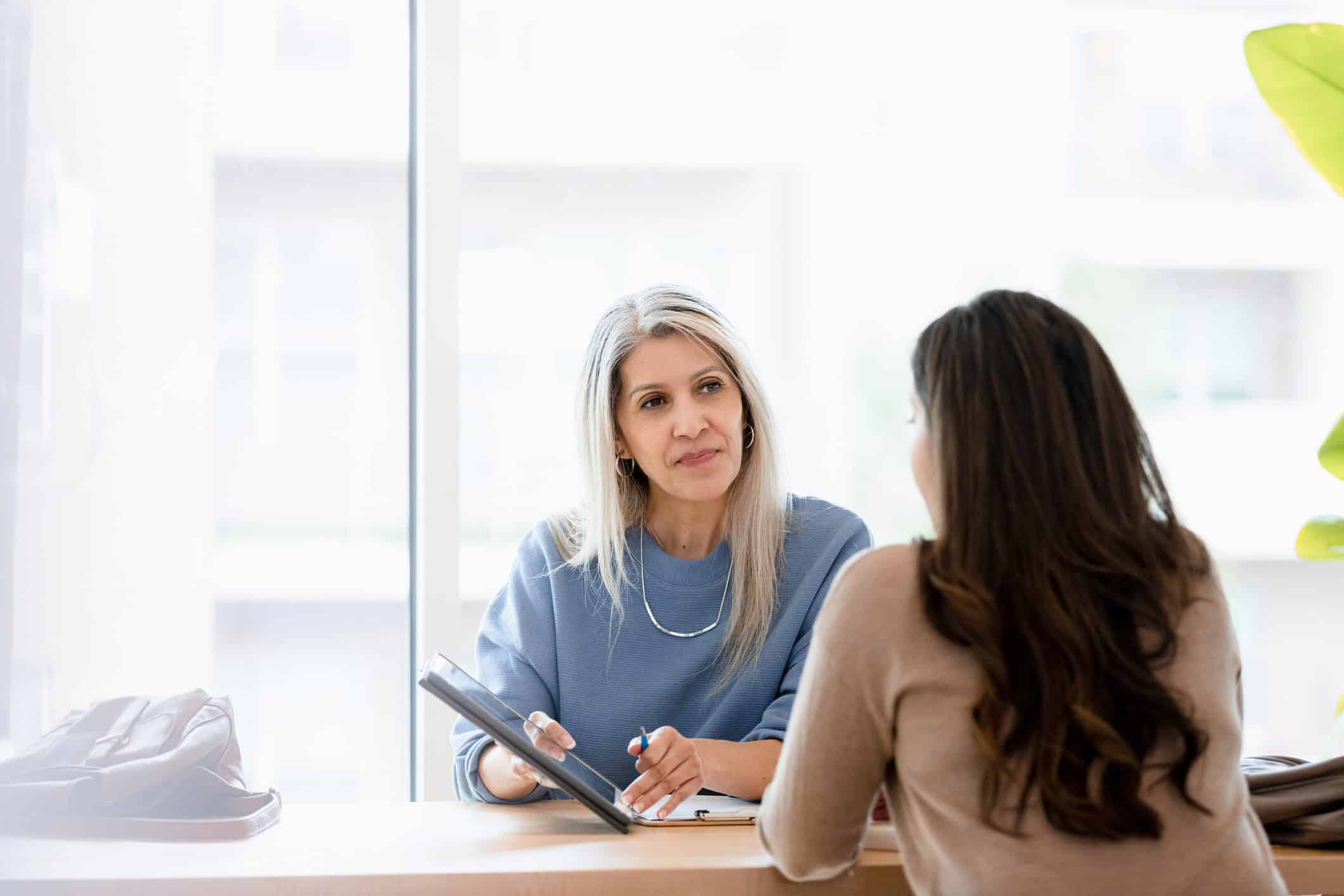 Two women discuss M&A Strategy Consulting Services at a table by a bright window.
