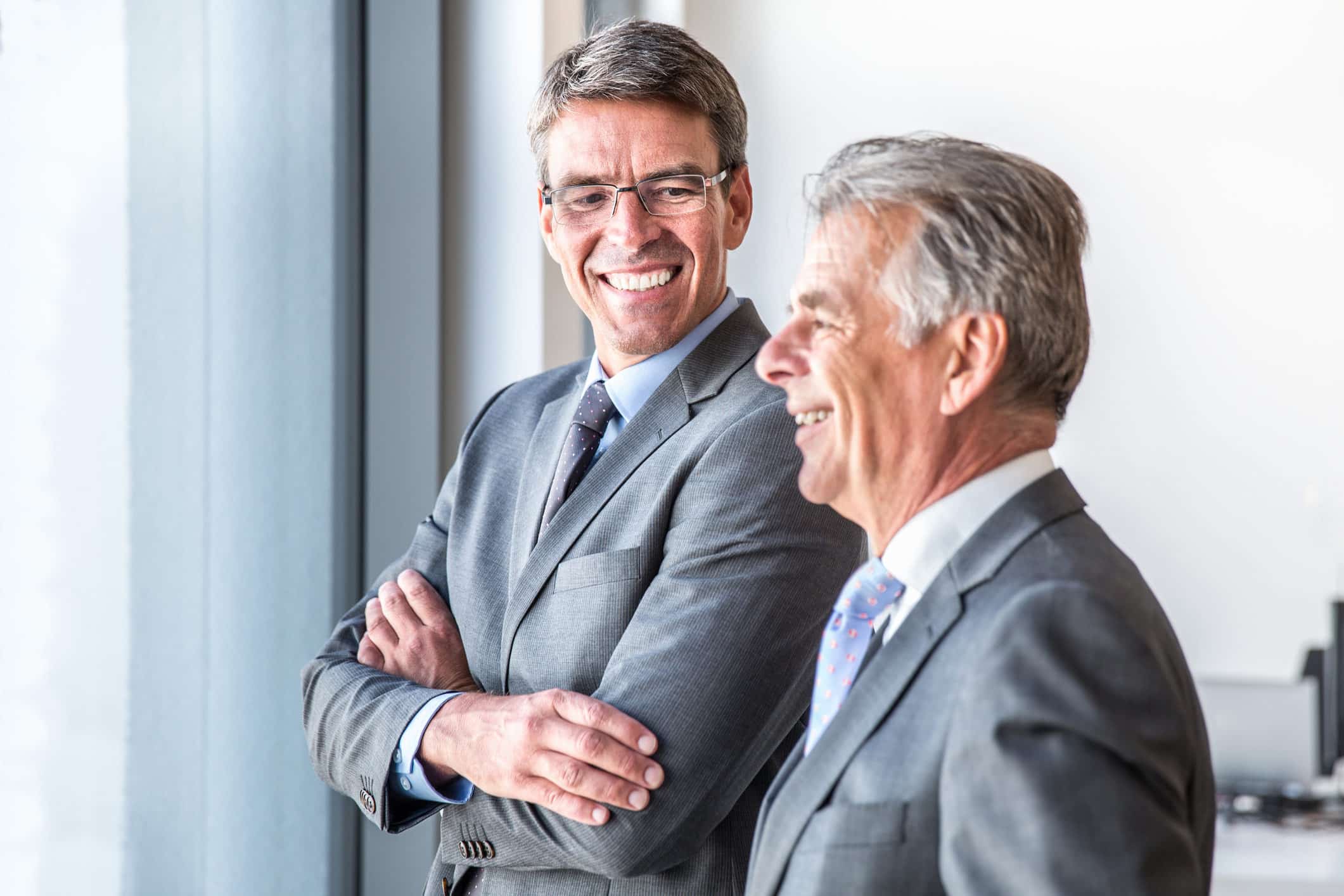 Two men in suits smiling and discussing forensic litigation by a window in a bright office.