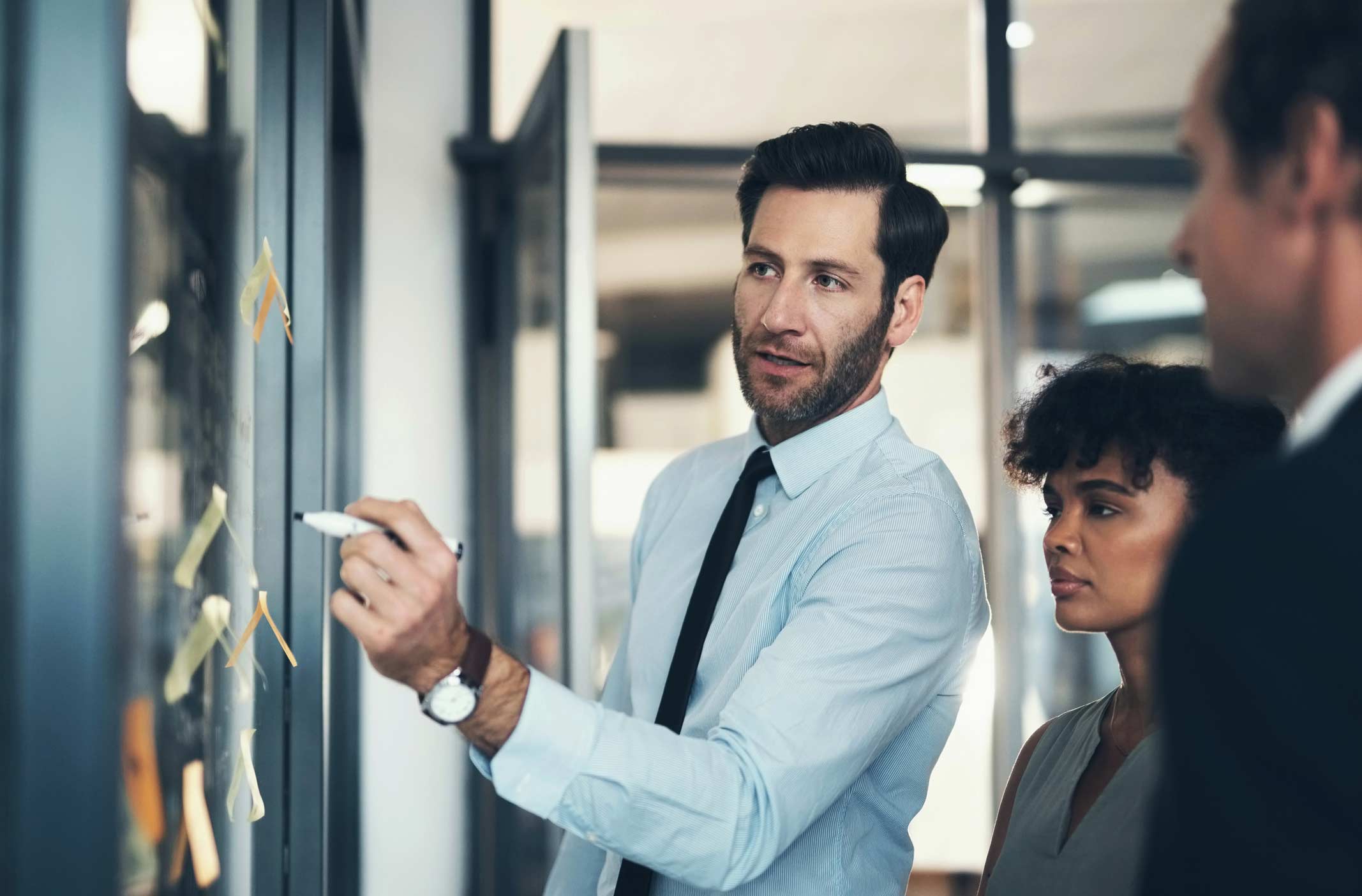 Man in a shirt and tie explains advanced analytics on a glass board as two colleagues watch.