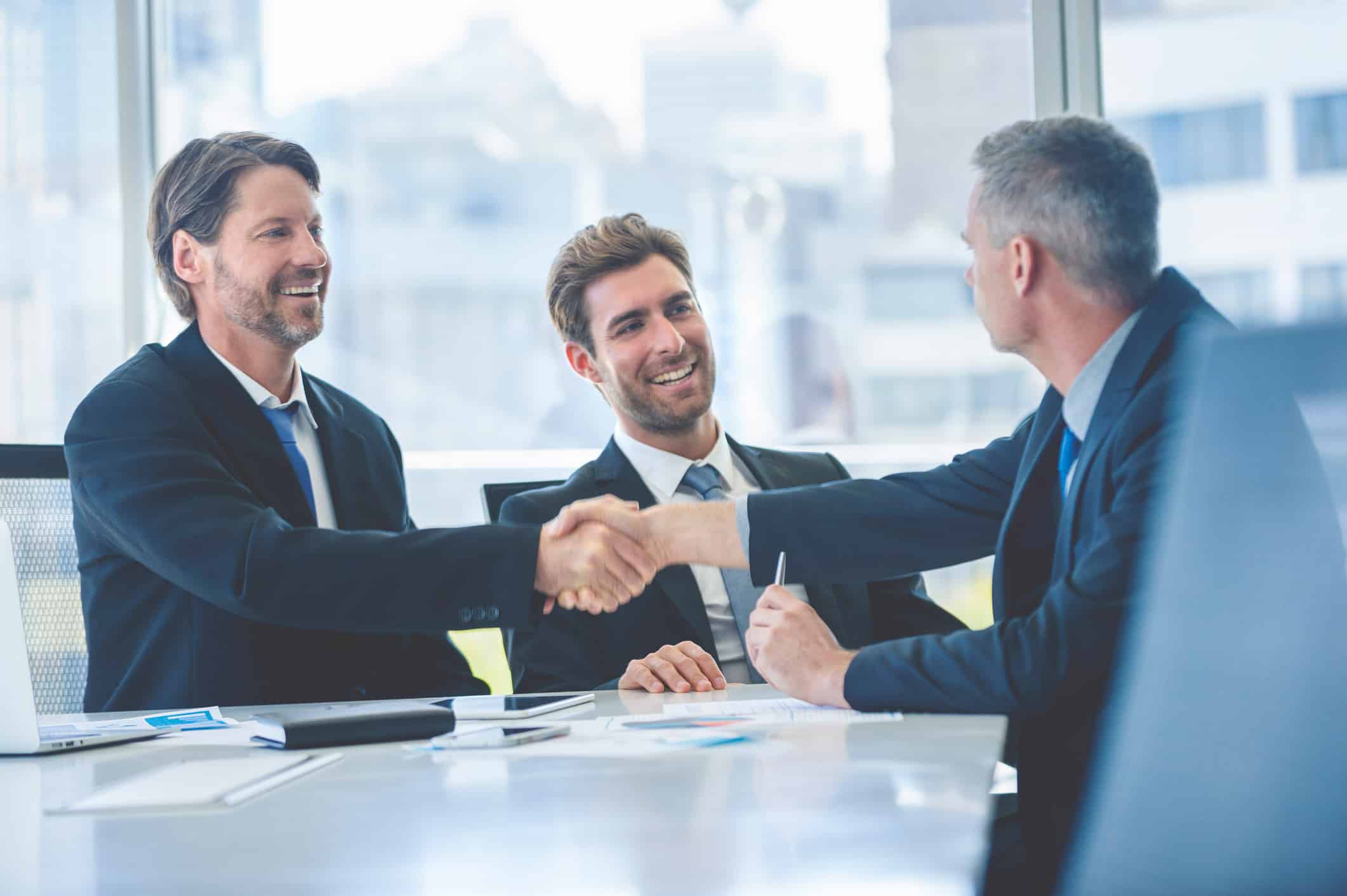 Three businessmen in suits smiling, shaking hands at a strategic planning meeting in an office.