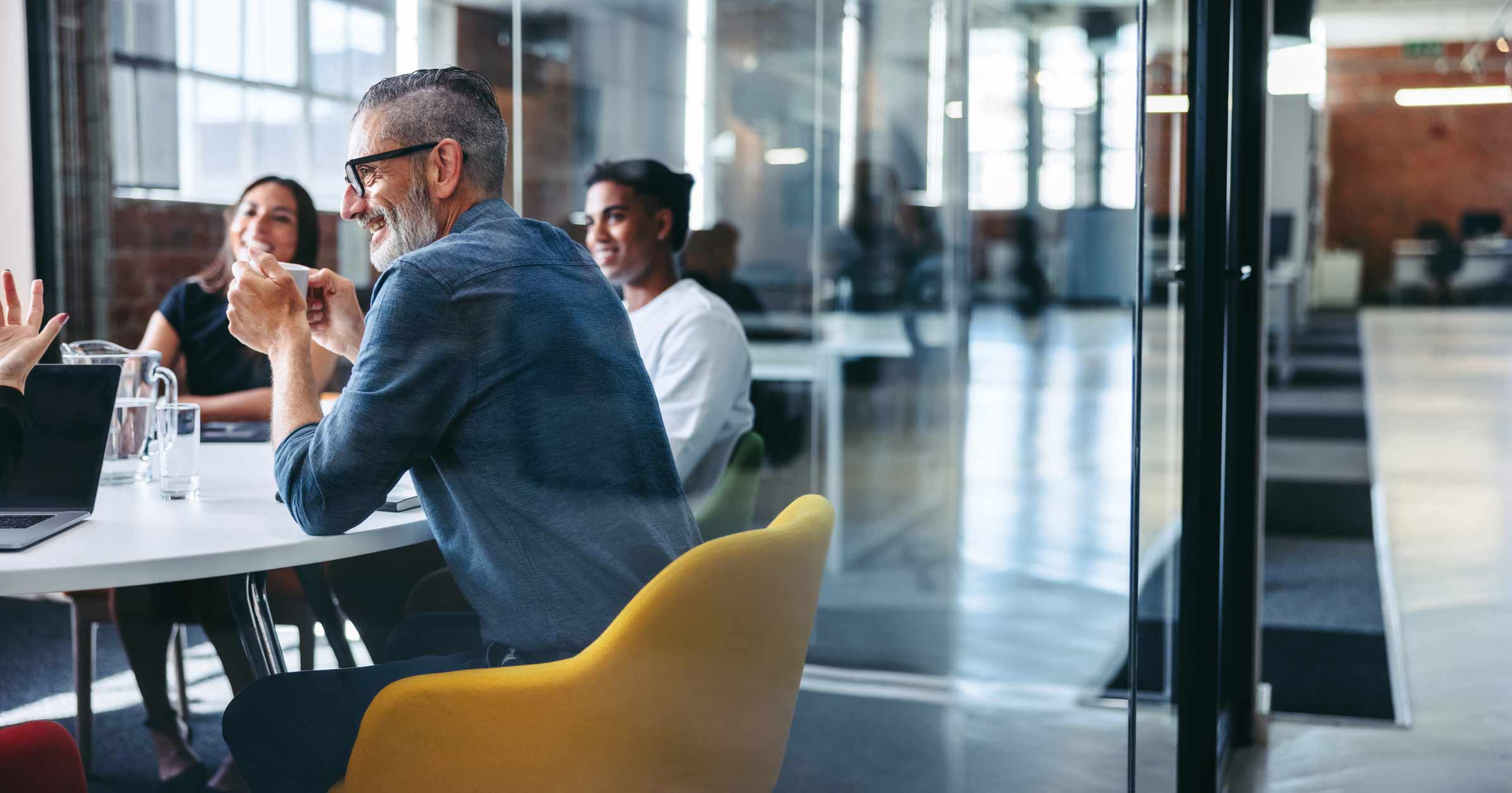 Four people sit at a table in a modern office, talking and smiling during a meeting.