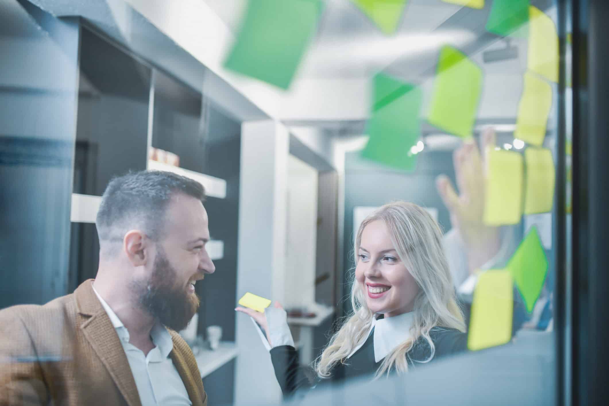 Two coworkers smiling and brainstorming Law Firm Pricing Architecture with sticky notes on glass.