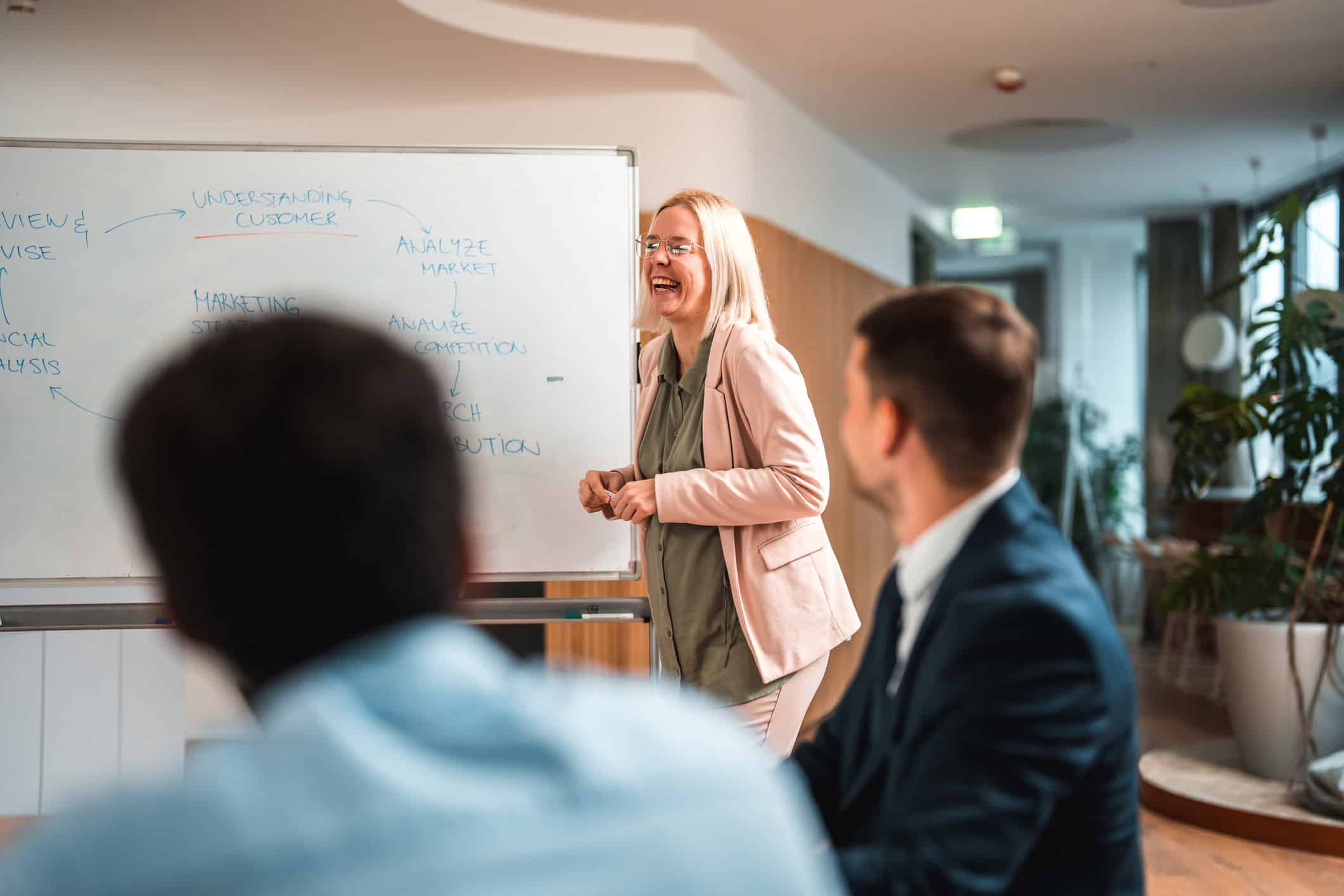 Smiling woman leads an M&A Integration Alignment discussion by a whiteboard in a modern office.