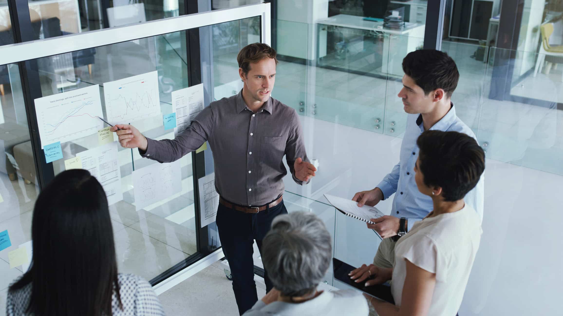 Man presenting charts on Talent & Resource Optimization to colleagues in a modern office meeting.