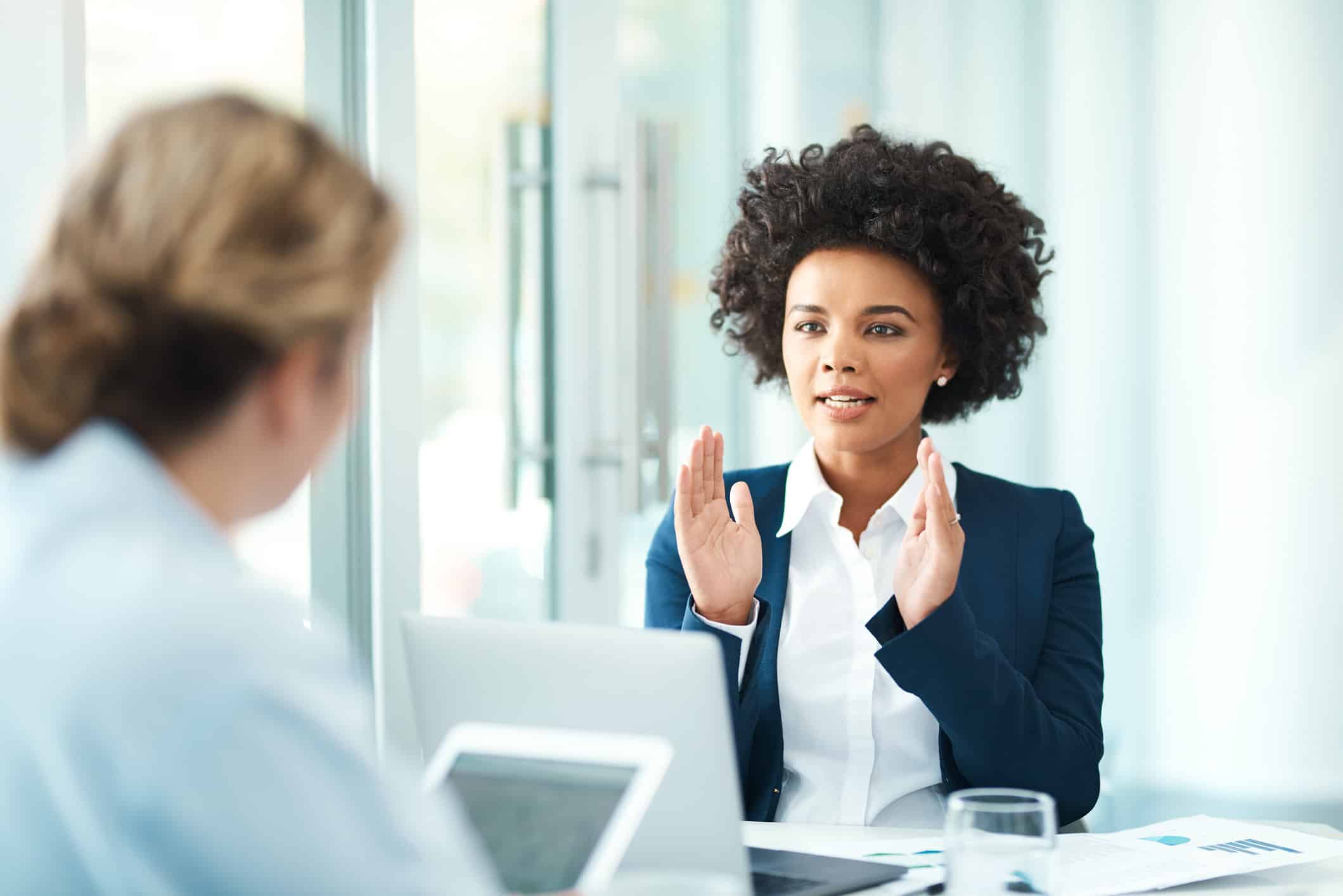 Two women discuss Business Intelligence Enablement across a table with laptops in a meeting.