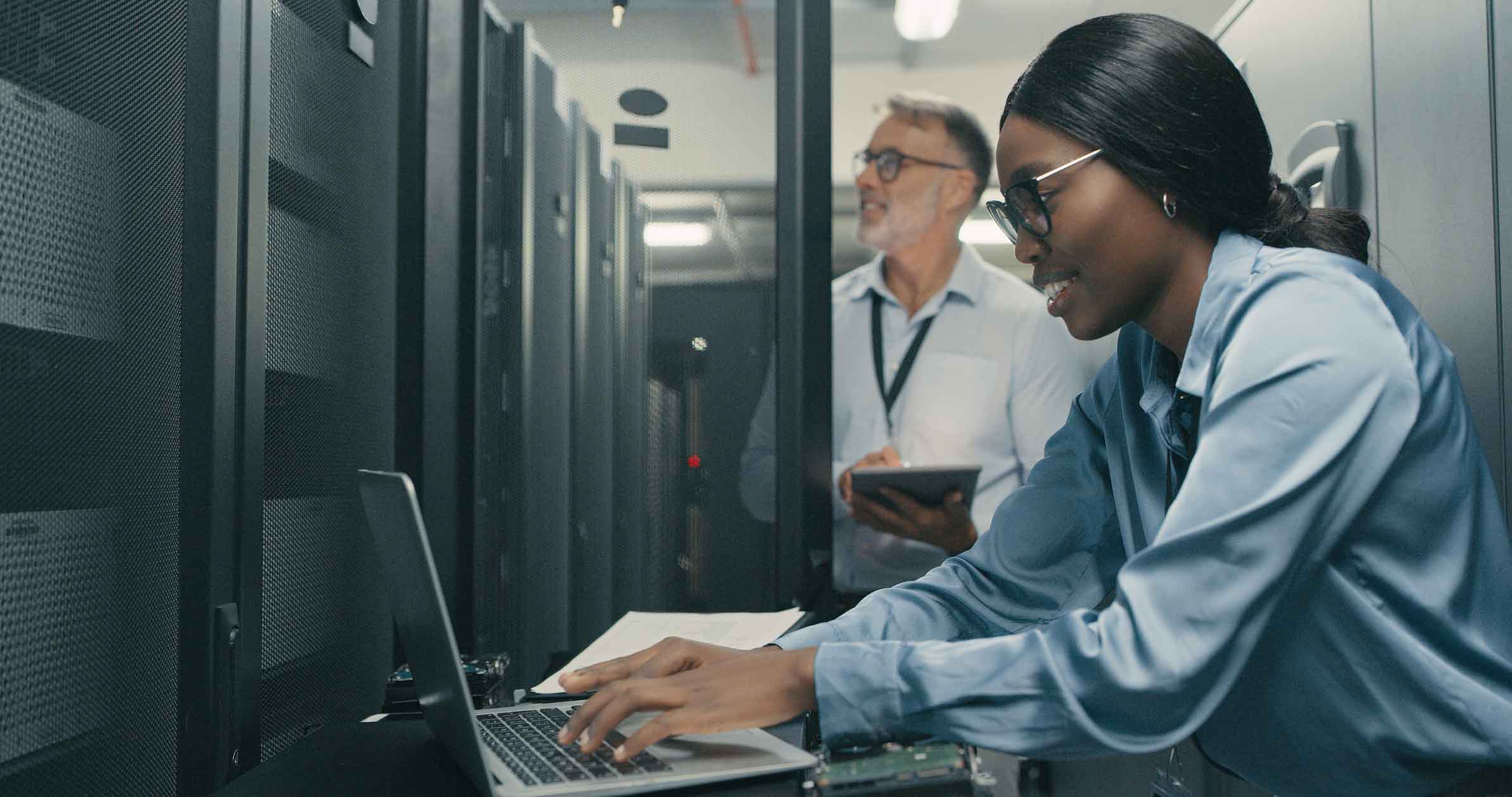 Woman working on laptop in server room, enabling information security; man with tablet behind.