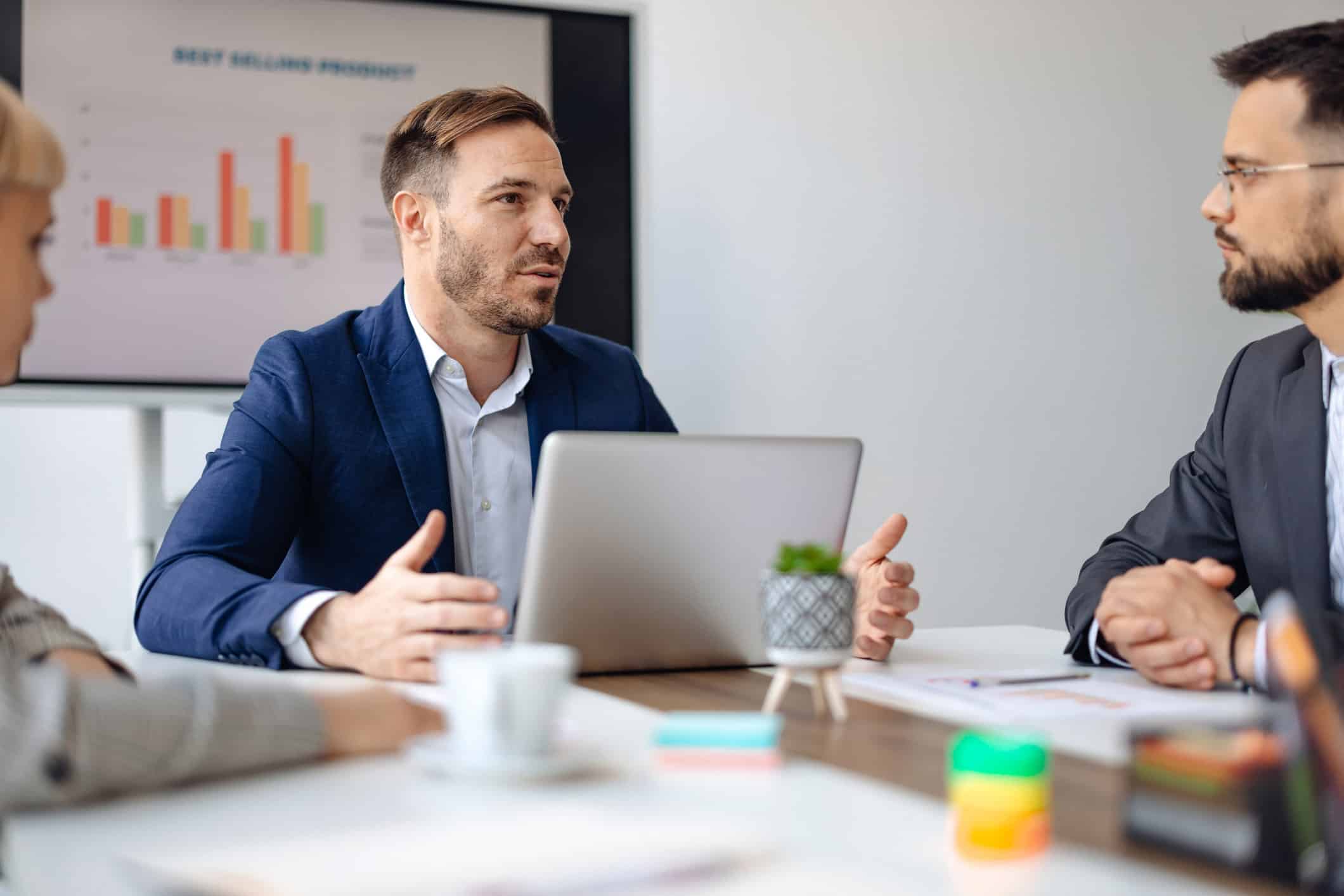 Man in suit speaks at meeting in a consulting firm, with laptop and chart in background.