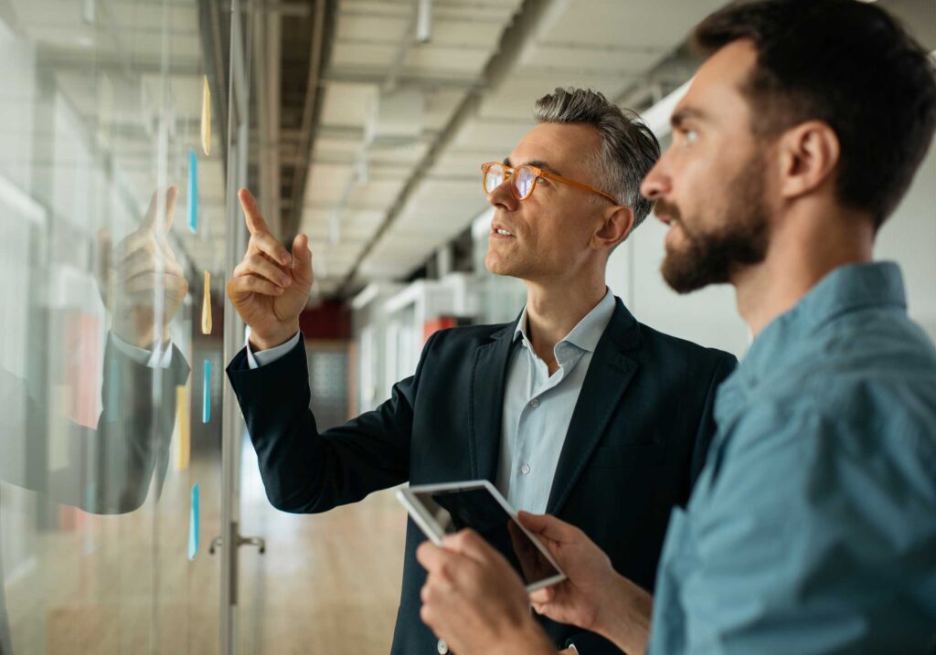 Two men discuss eDiscovery notes on a glass wall; one holds a tablet, consulting together.
