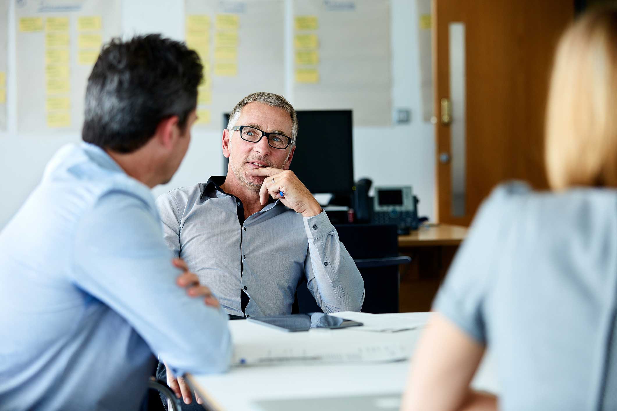 Three people discuss identity access management consulting in an office, papers on the table.