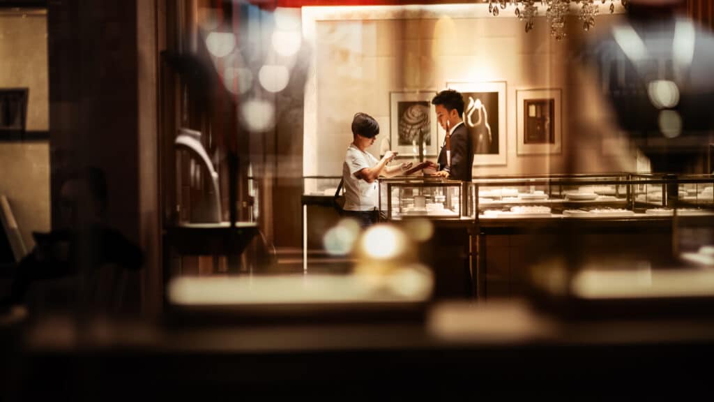 Two people talking at a jewelry store display counter, with warm, soft lighting.