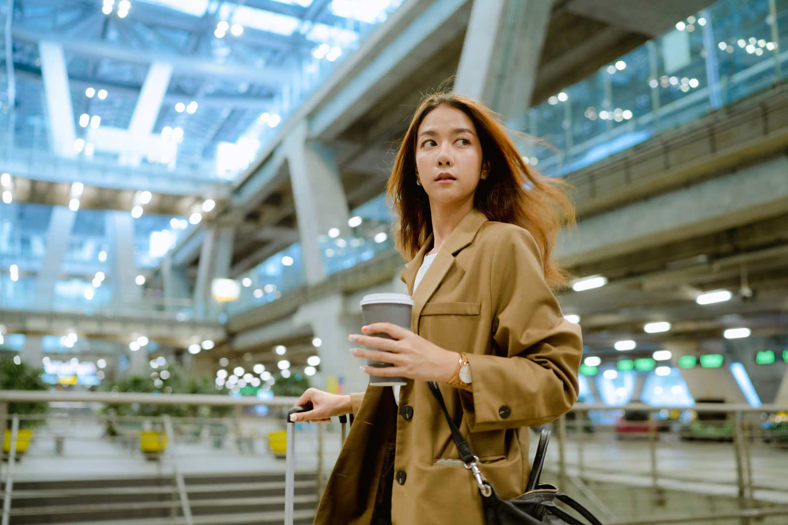 Woman in a brown coat holding coffee and luggage, pondering Retention Analytics in an airport.