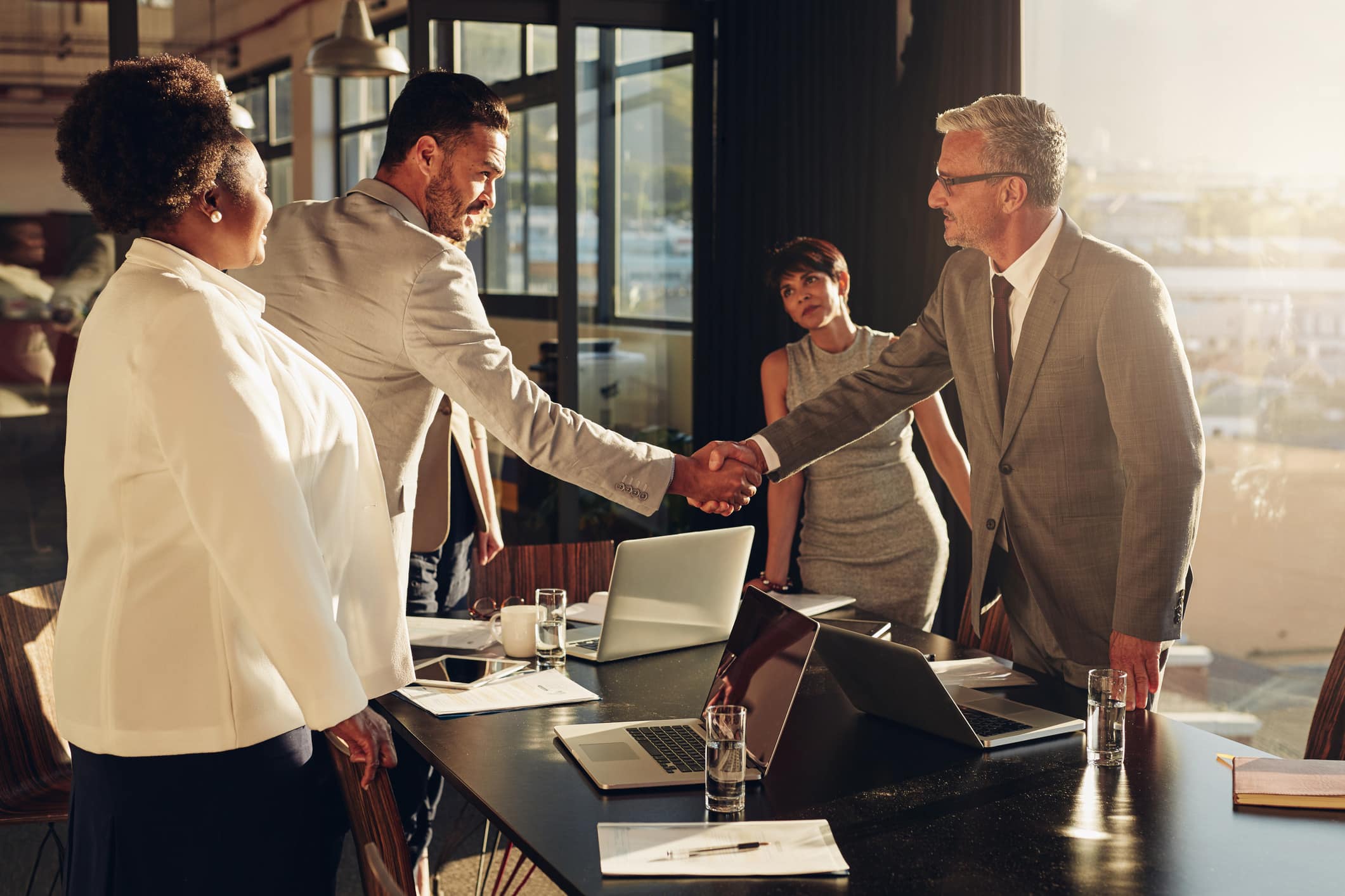 Four businesspeople in a meeting room discuss M&A Due Diligence Consulting, two shake hands.