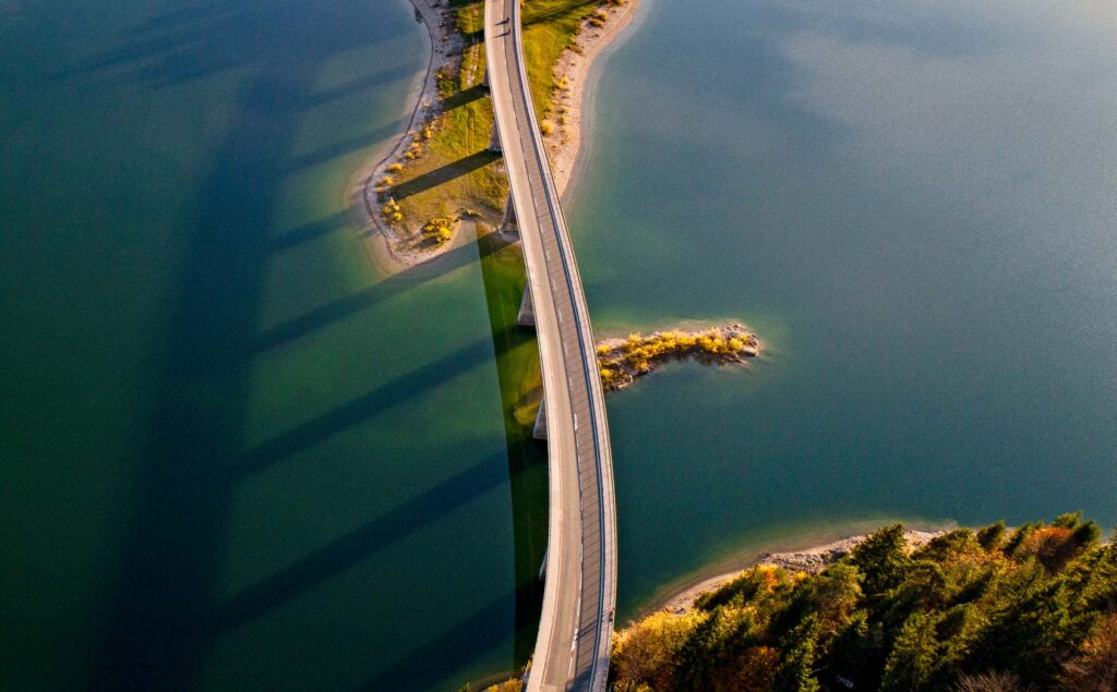 Curving bridge casts long shadows near green-blue water, connecting to aviation consulting services.
