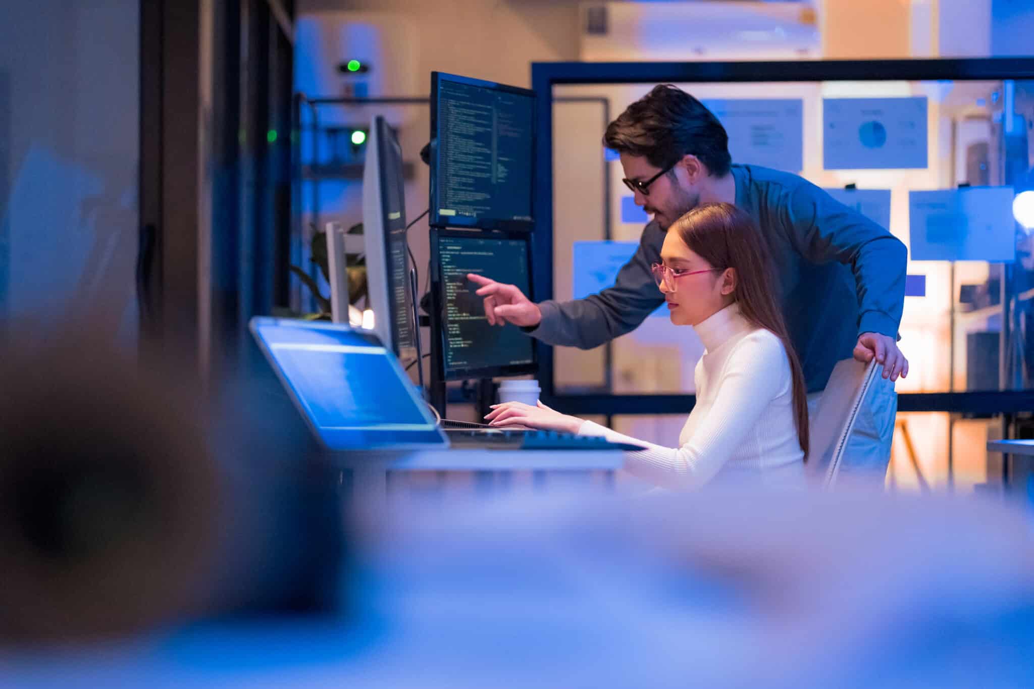 Two people working together at computers with code on the screens in a modern office.