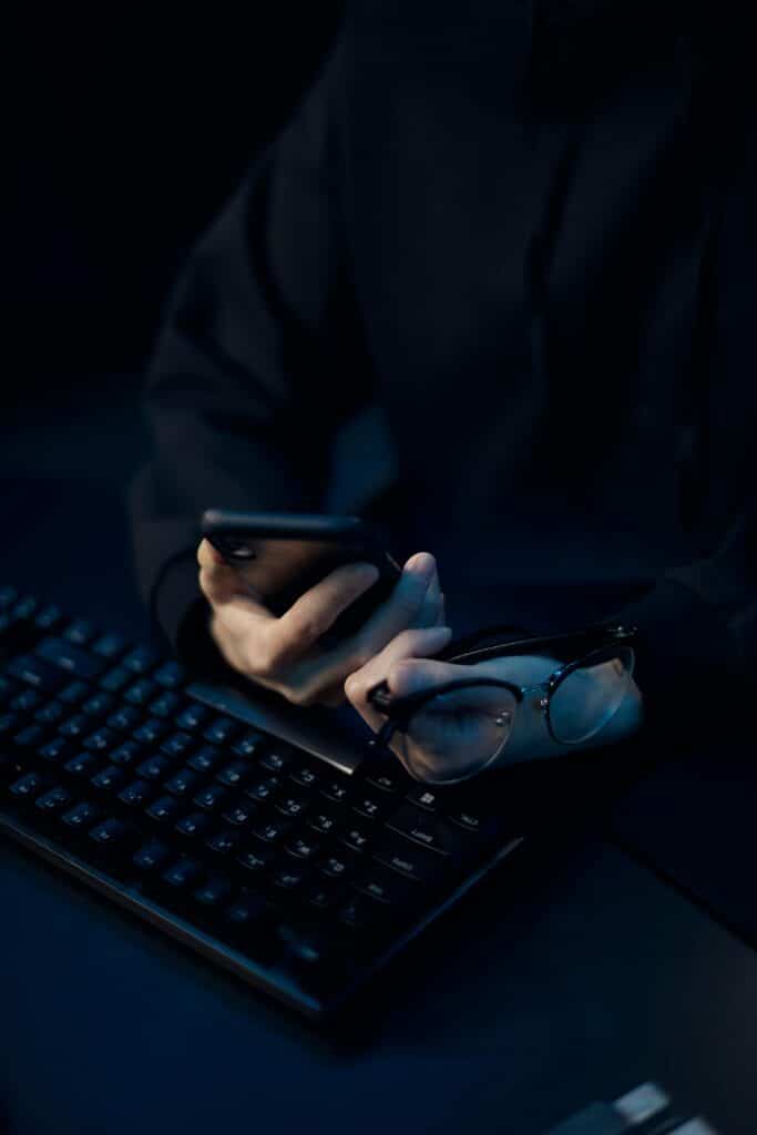 Person holding a phone and eyeglasses near a computer keyboard in low light.