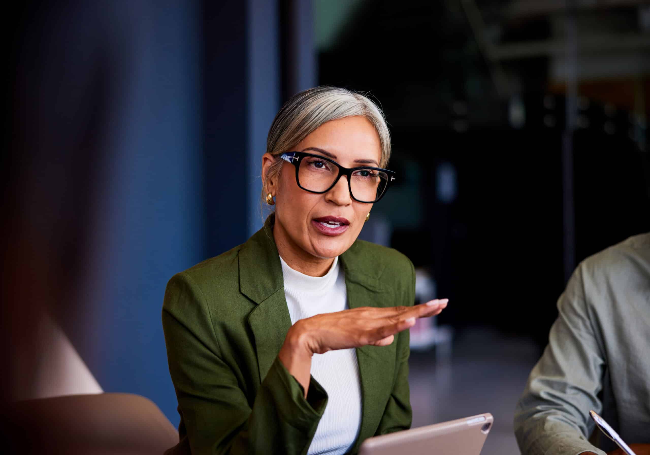Woman with gray hair and glasses, in green blazer, gesturing while discussing AI governance.