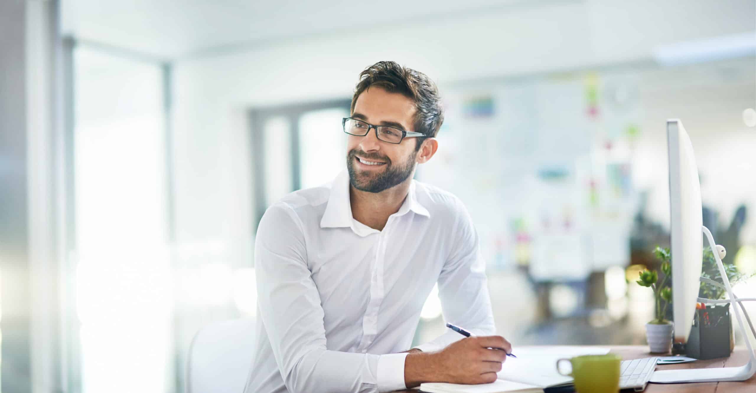 Man in glasses smiling at desk, writing notes for commercial real estate transaction management.