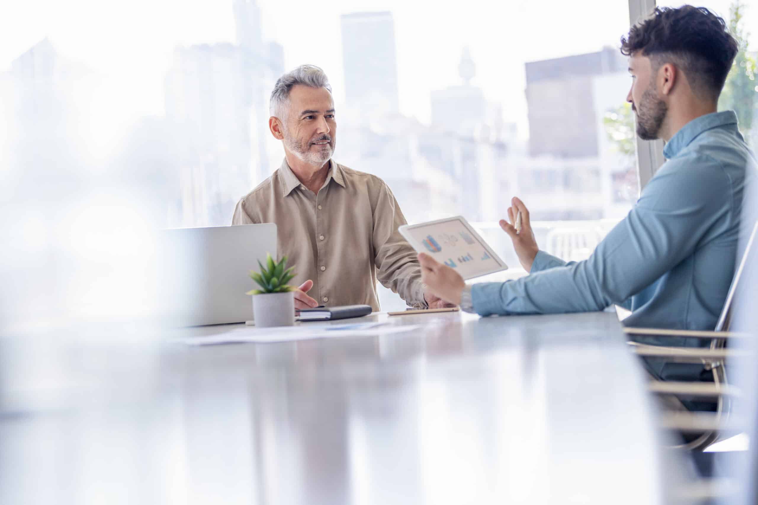 Two men in an office discuss cloud migration on a tablet at a conference table with a laptop.