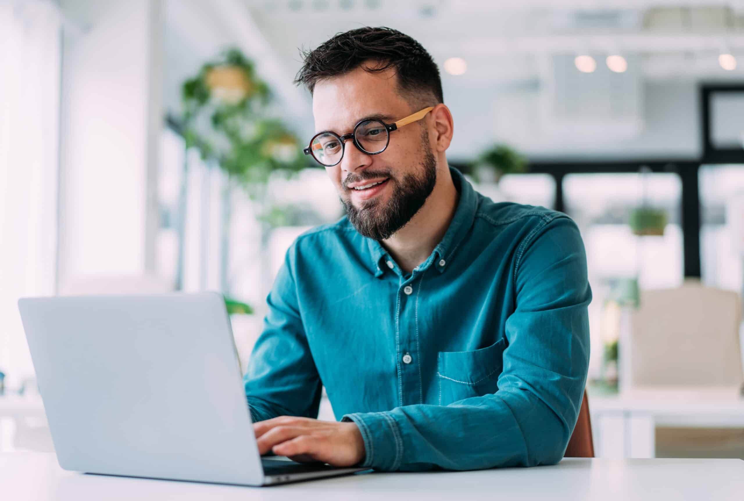Man with glasses smiling and working on a laptop in a bright digital twins consulting office.