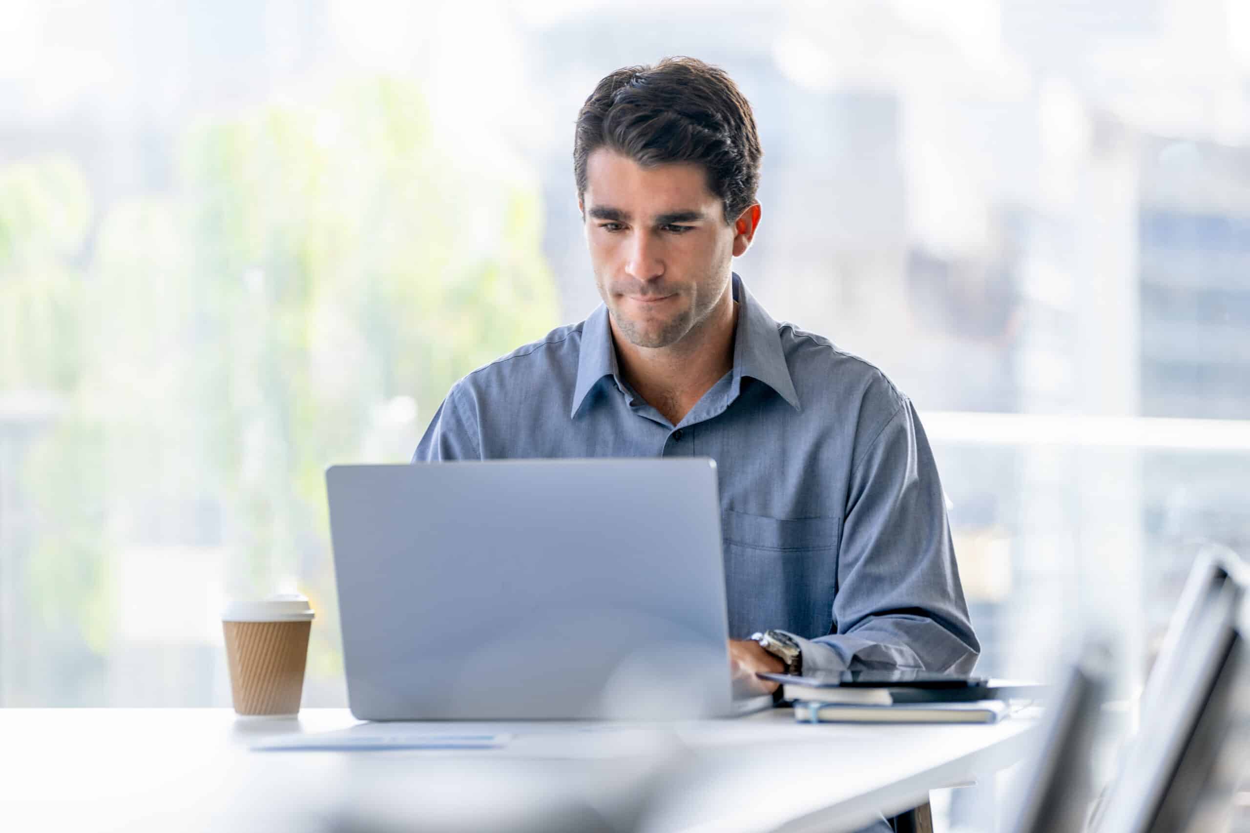 Man in a blue shirt works on enterprise systems transformation at his desk with coffee and a notebook.