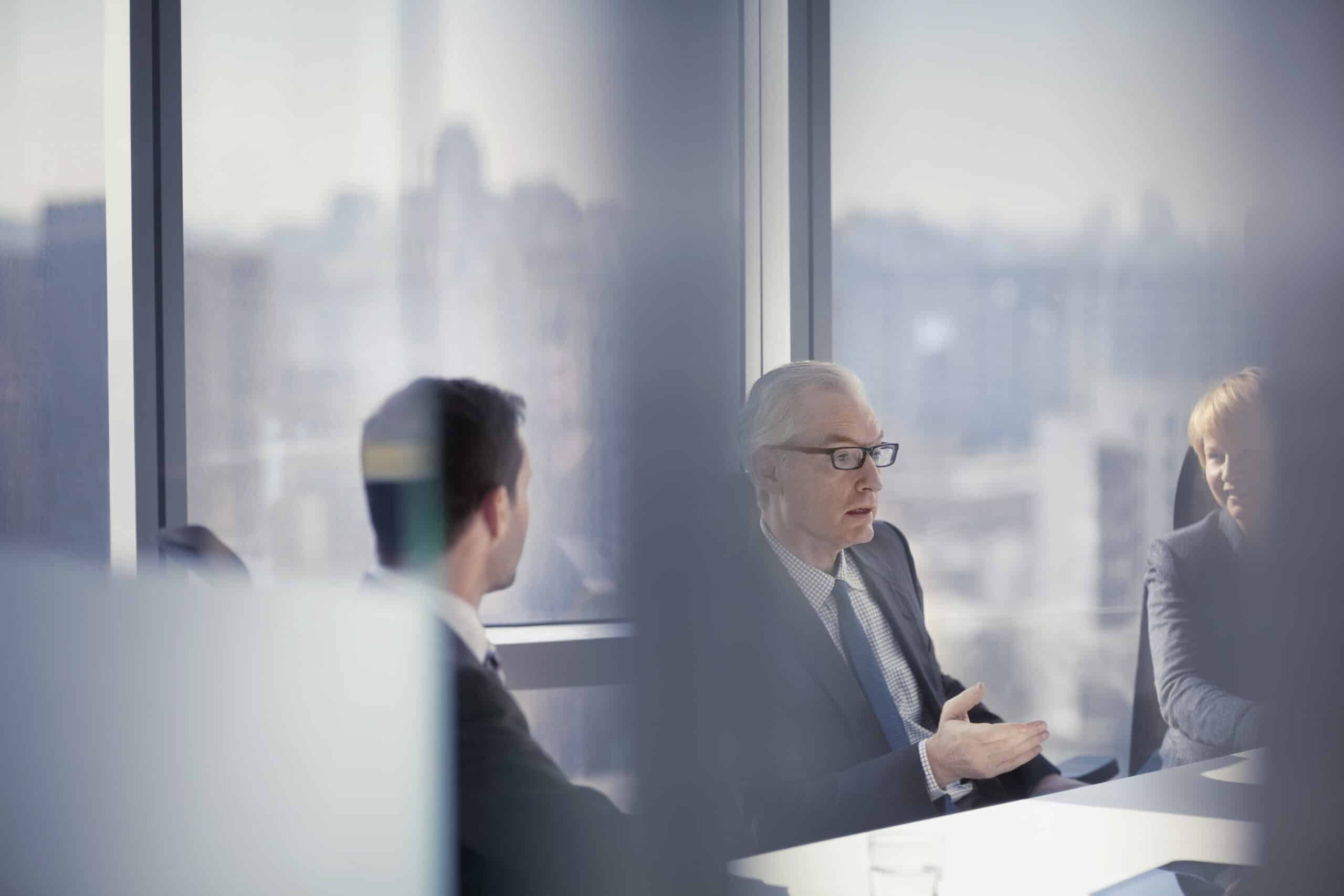 Three people discuss IT architecture consulting at a conference table by large windows.