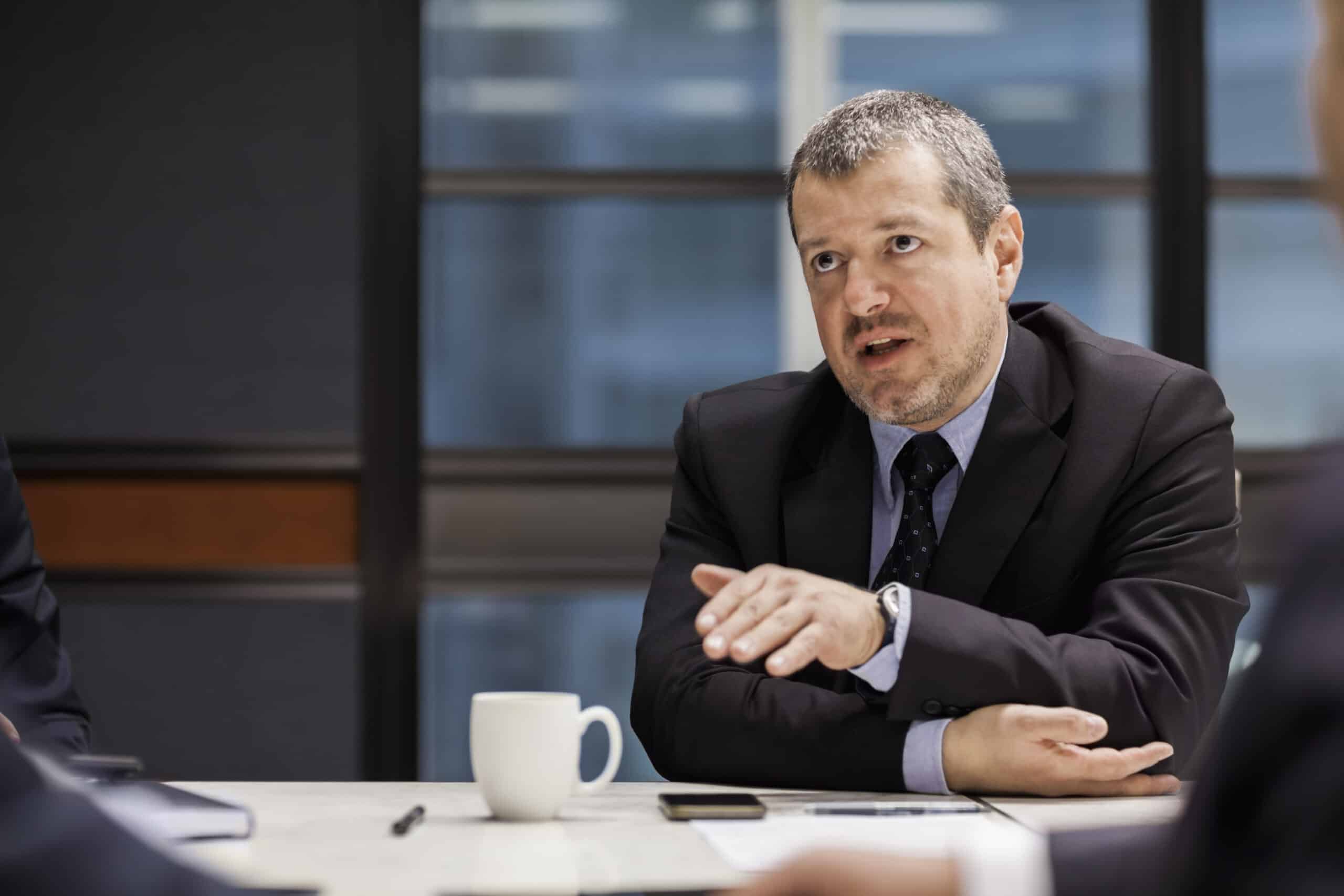 Man in suit discussing IT architecture consulting at a meeting, gesturing with hand.