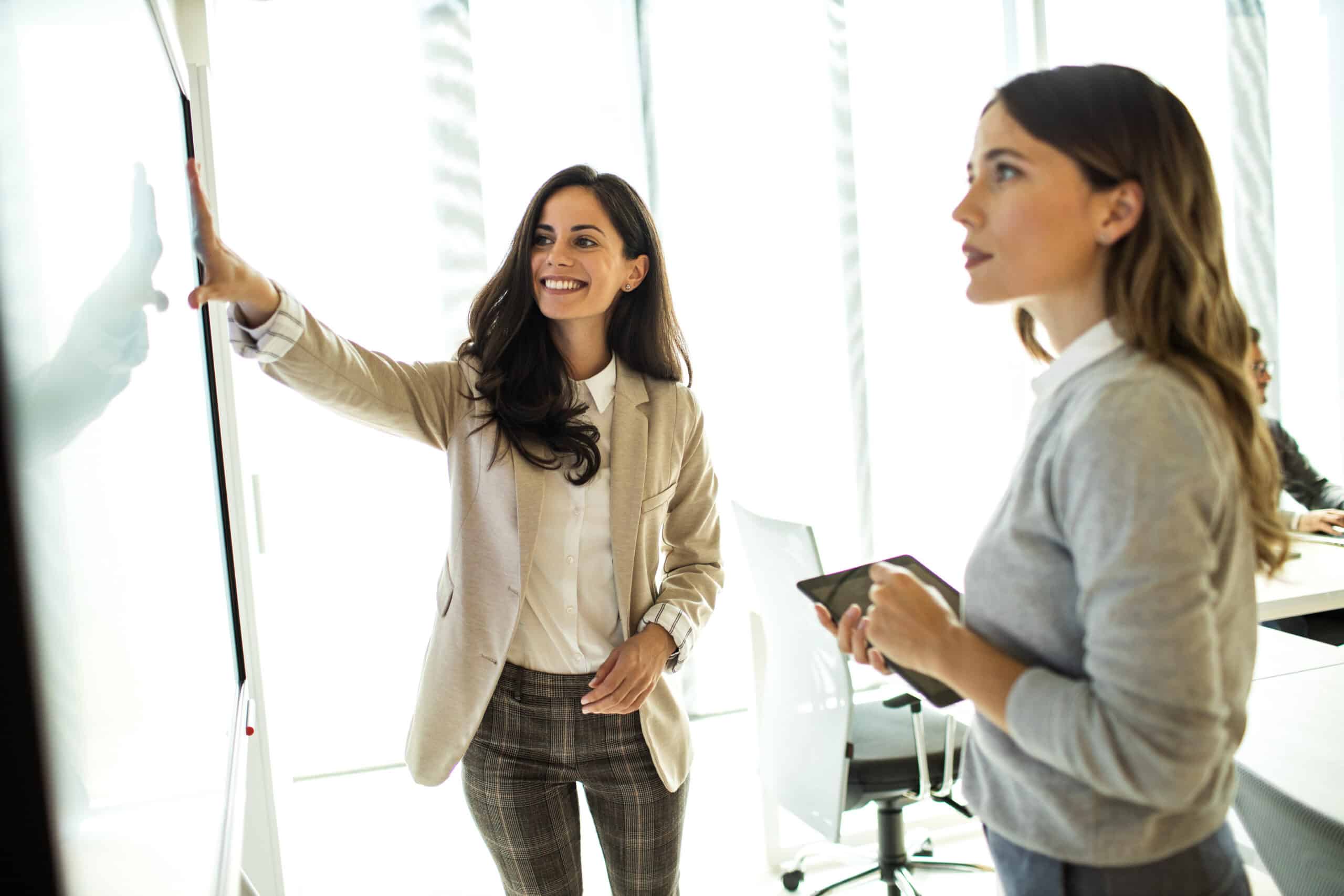 Two women discuss and point at a whiteboard in an it governance consulting meeting room.
