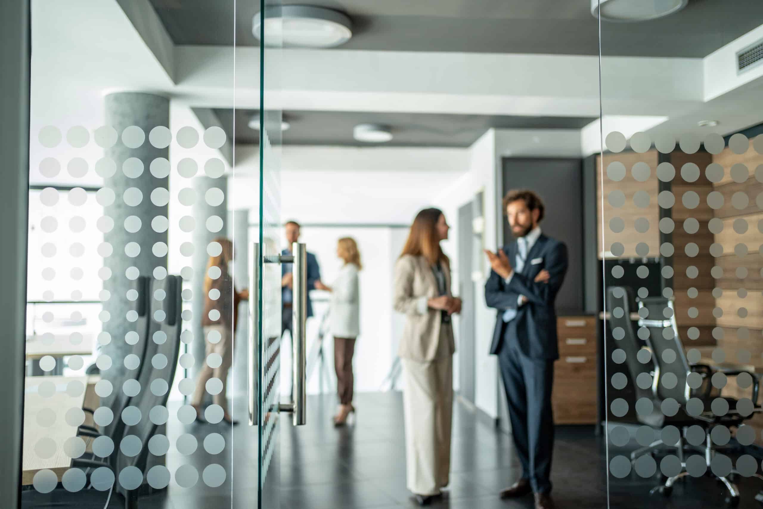 Four it governance consultants in business attire talk in a modern office behind a glass wall.
