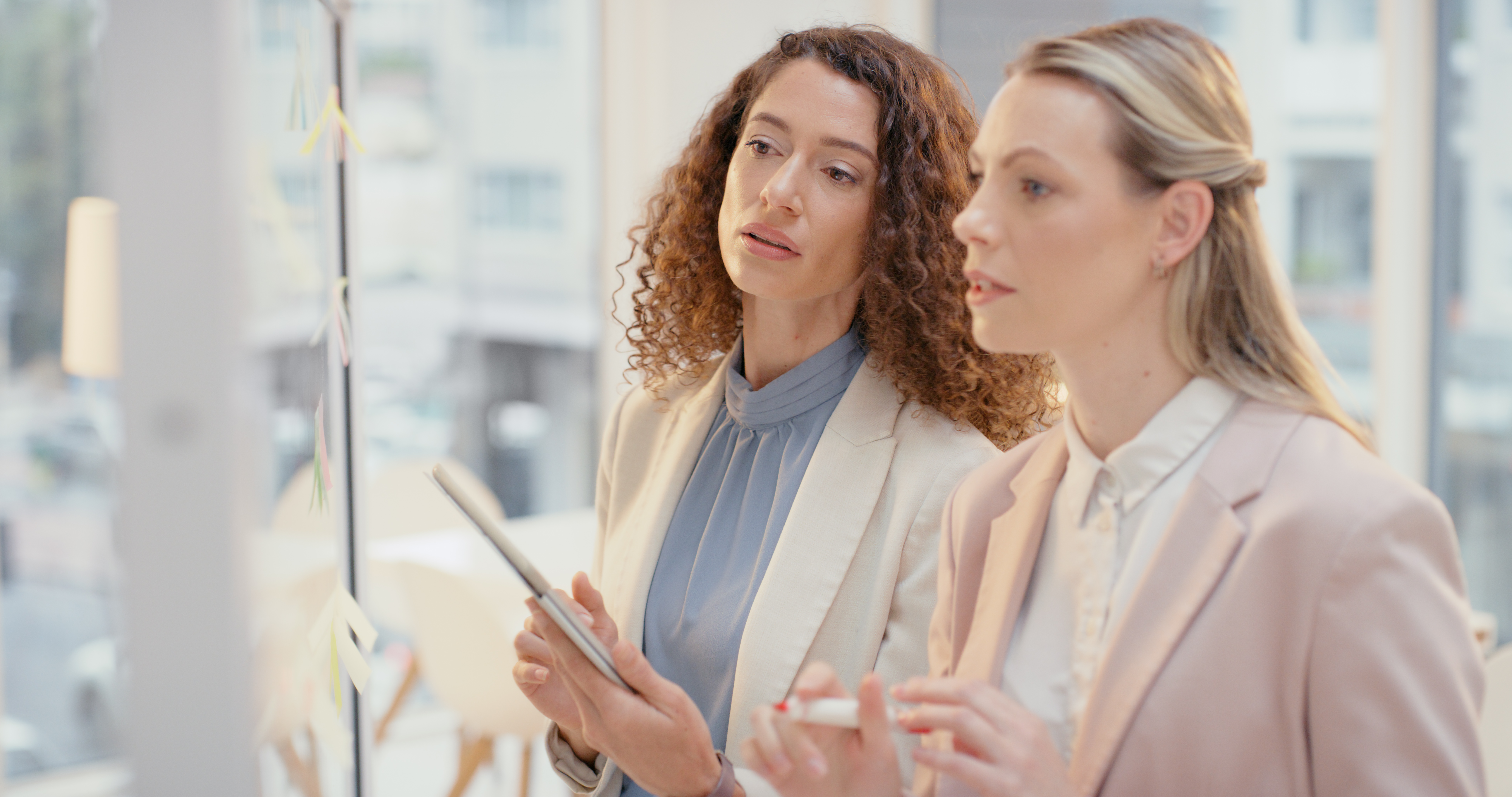 Two women in business attire discuss ideas and it governance on notes at a glass office wall.