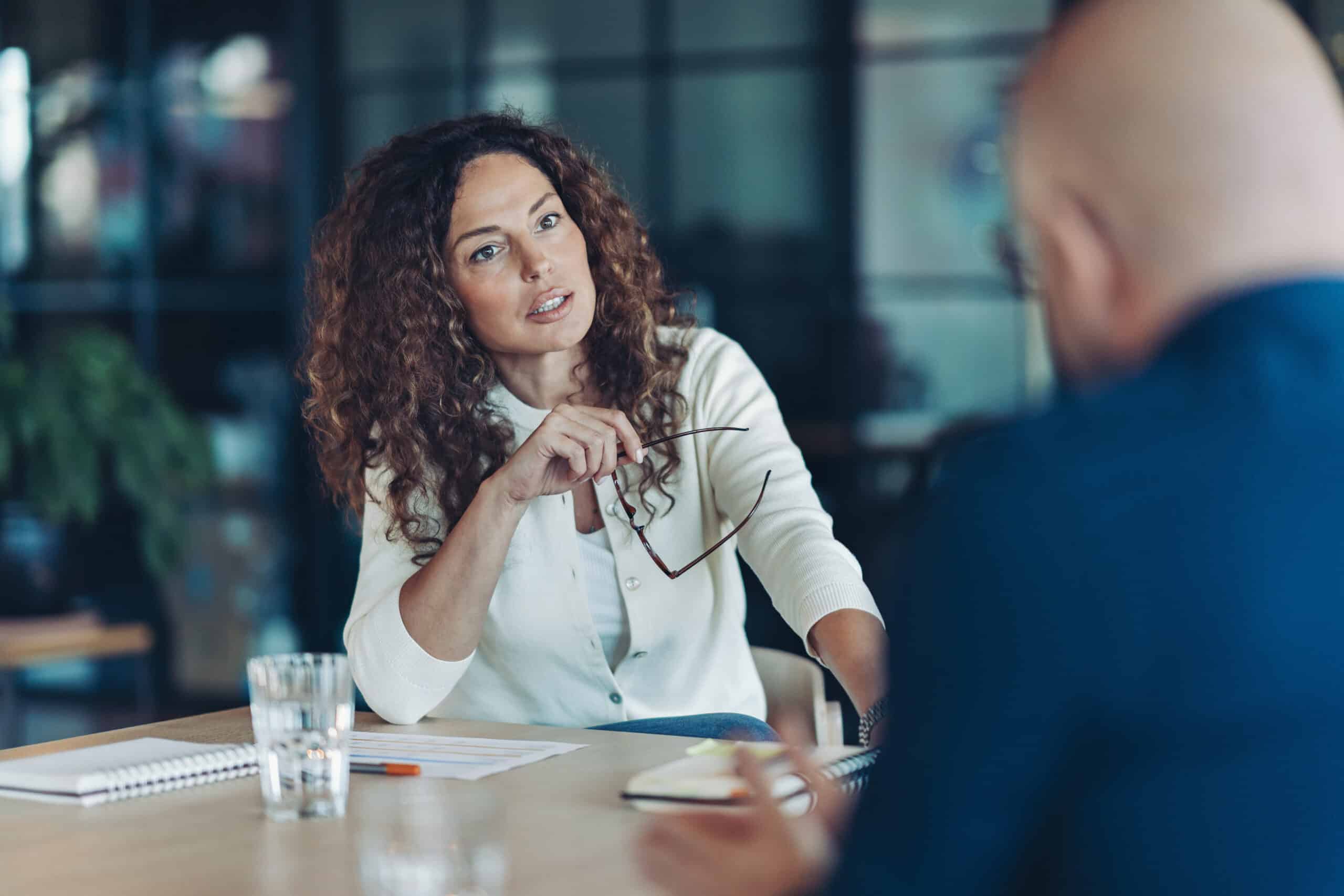 Woman holding glasses discusses it infrastructure consulting with a colleague at a table.