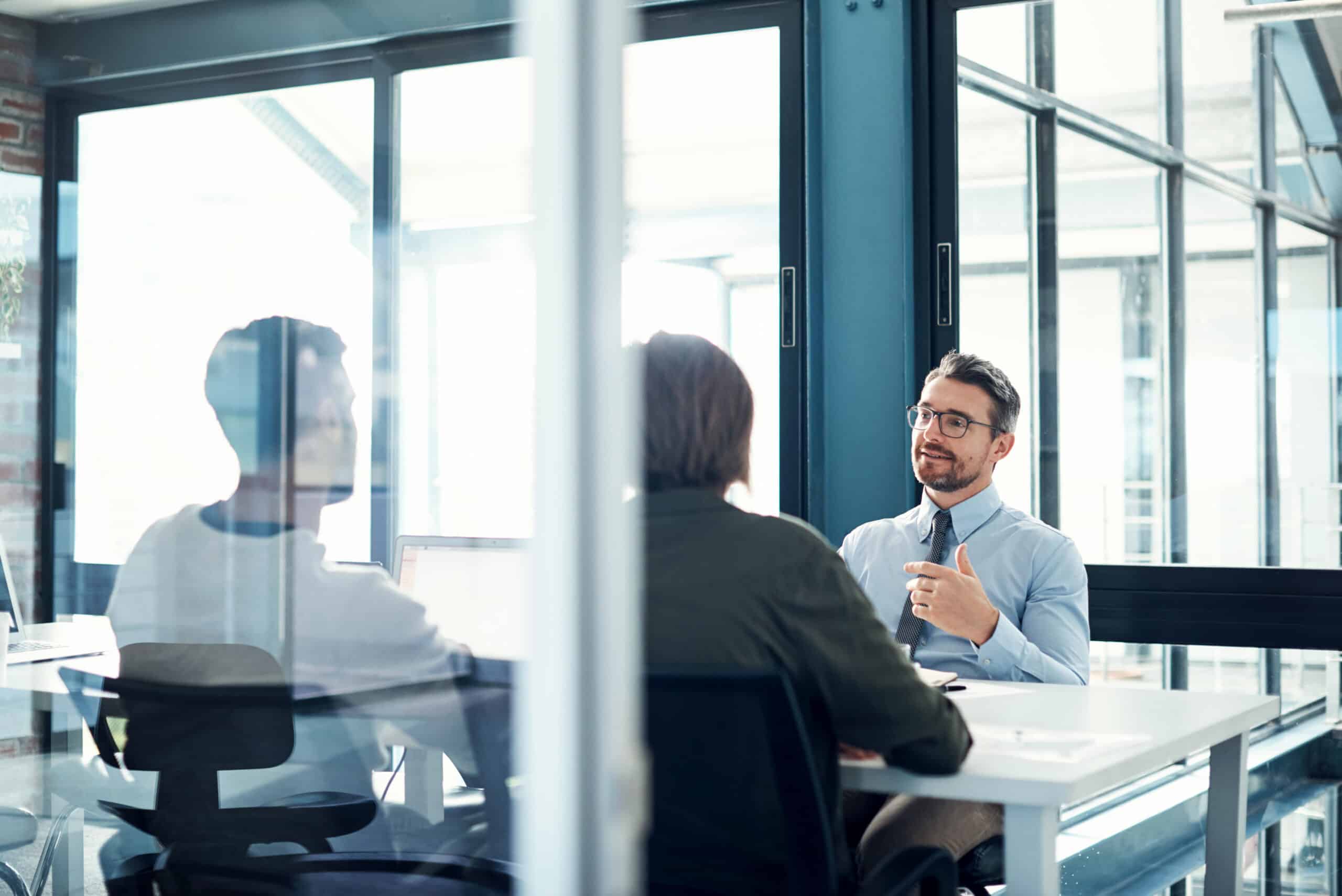 Three people discuss IoT consulting in a modern office with large glass windows.