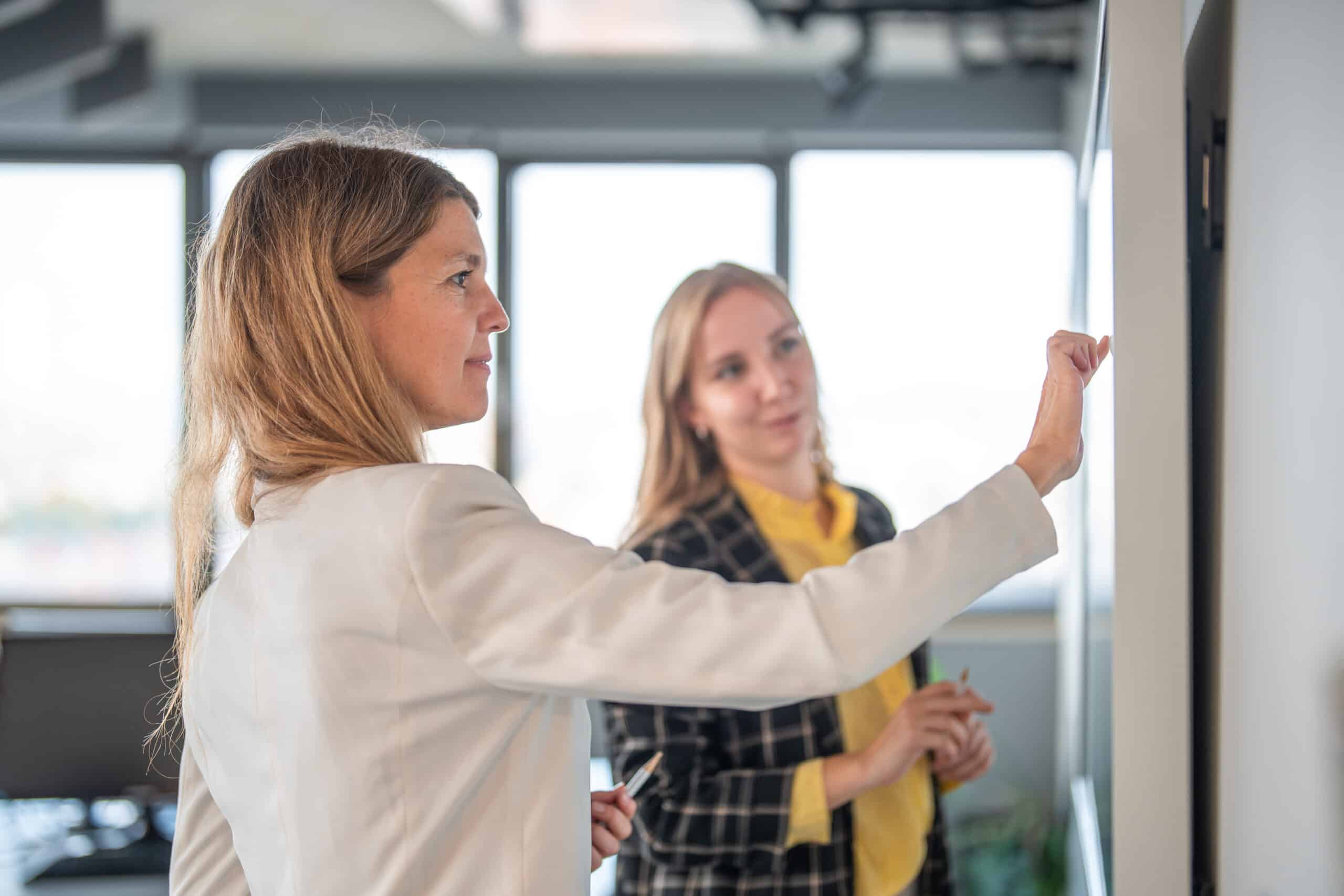 Two women discussing IoT solutions and writing on a whiteboard in a bright office setting.