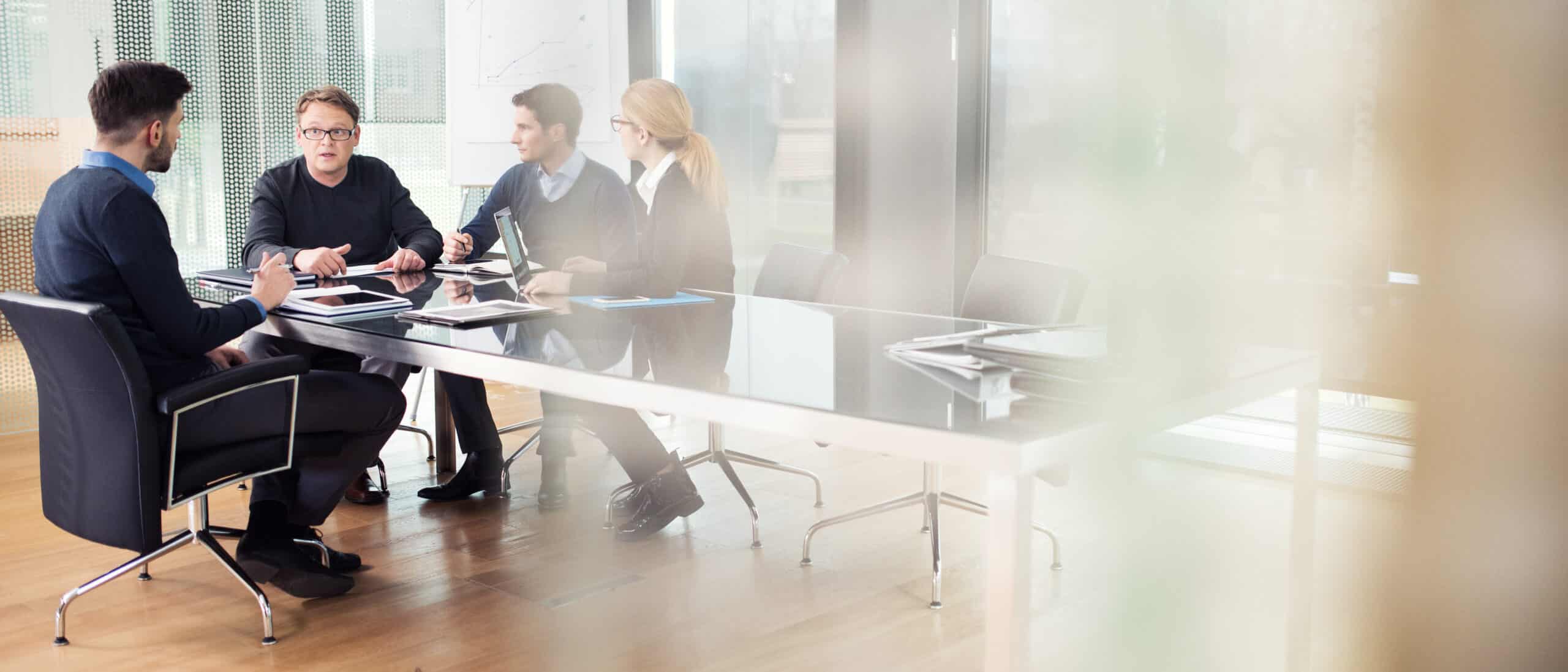 Four IoT consultants in business attire meet around a glass conference table in a modern office.