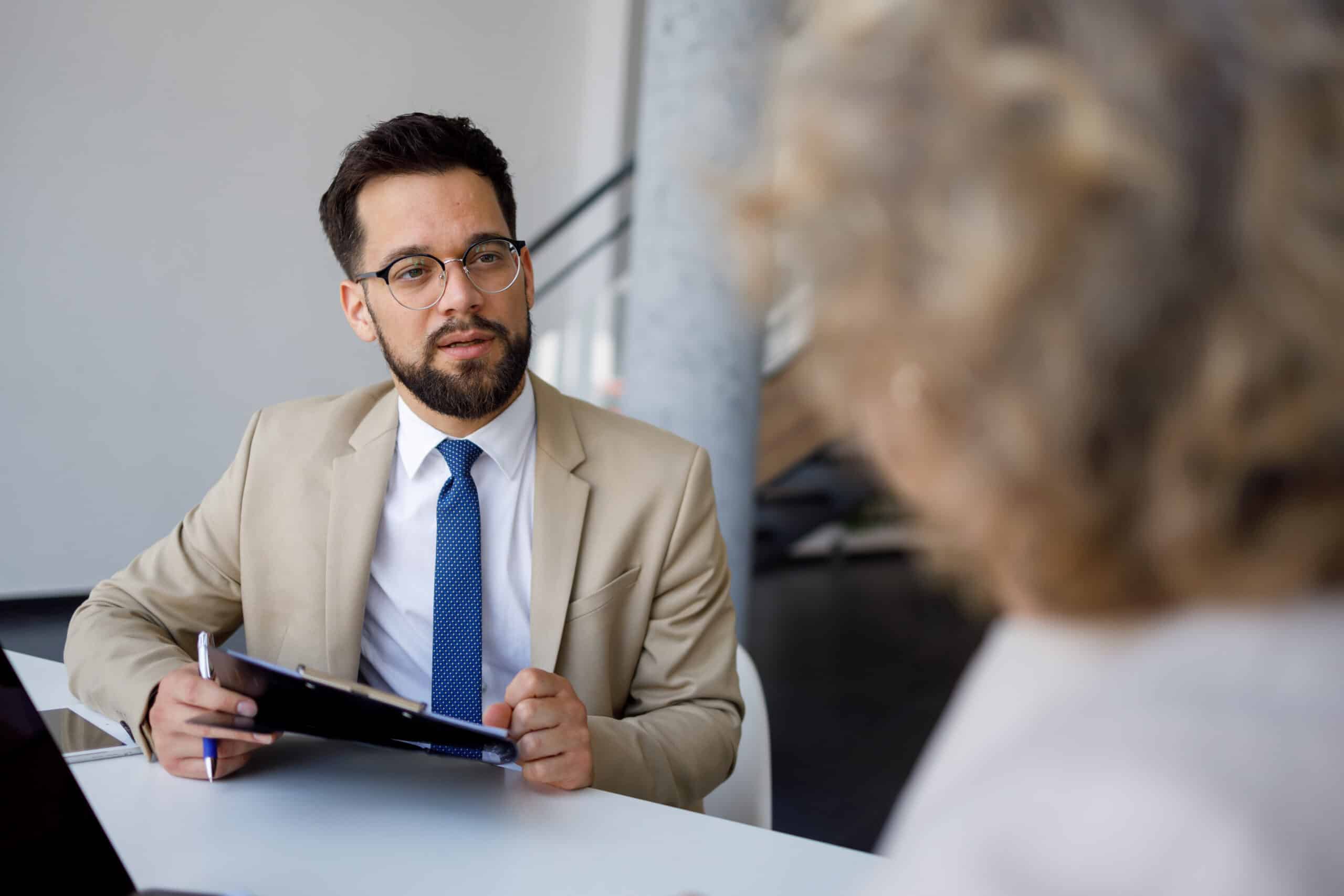 Man in suit holding clipboard, discussing leadership development with a woman at her desk.