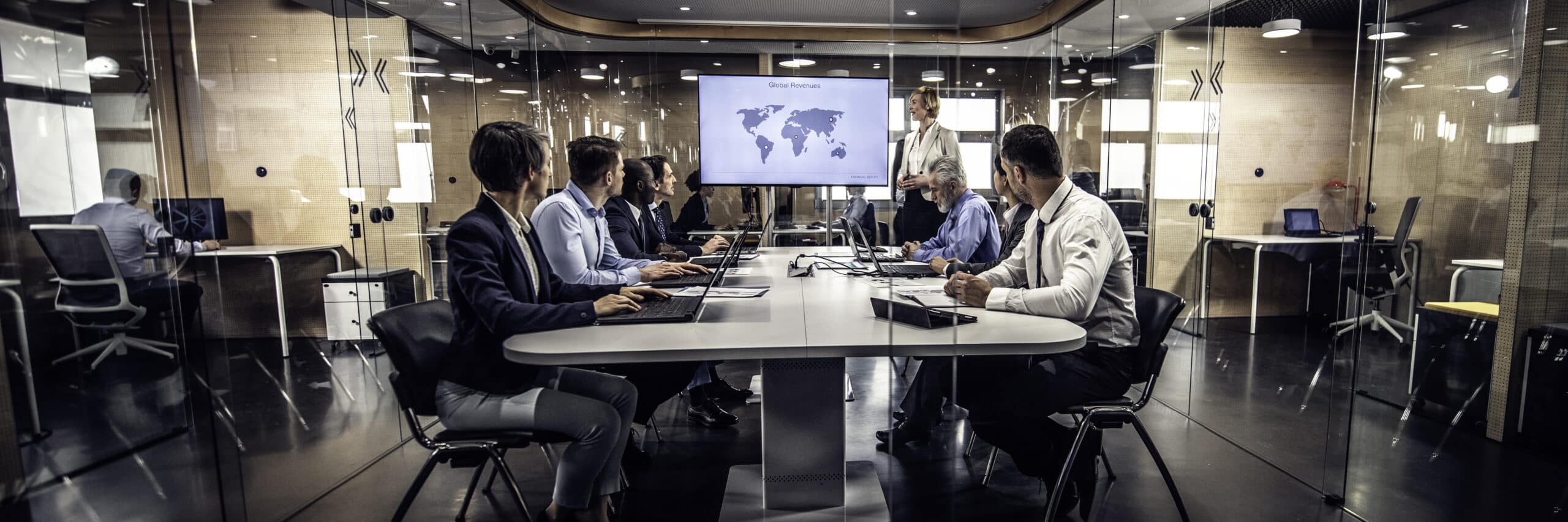 Seven people in a modern glass office discuss location intelligence consulting with a world map onscreen.
