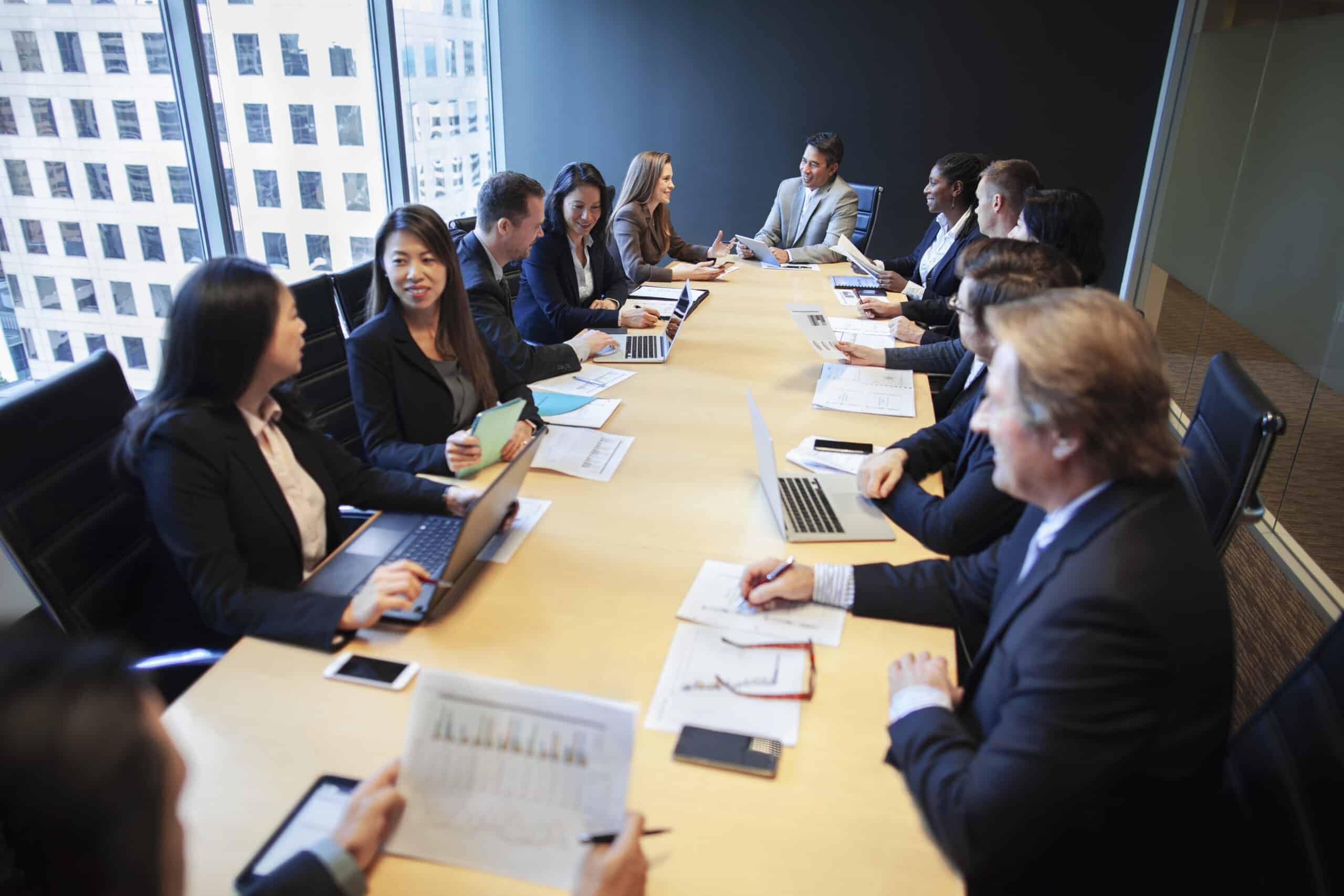 People in business attire discuss NLP consulting around a conference table with laptops and papers.