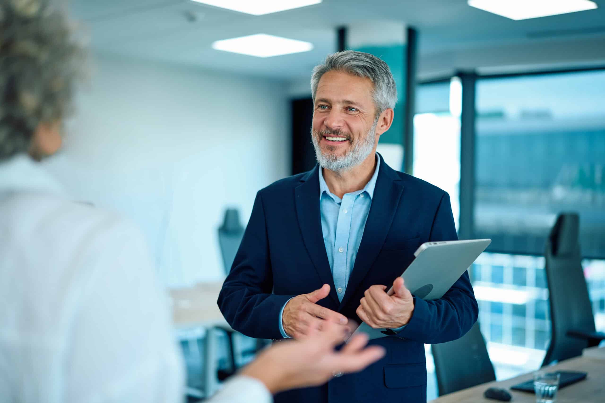 Smiling man in a suit holding a tablet, discussing proptech strategy with a colleague in an office.