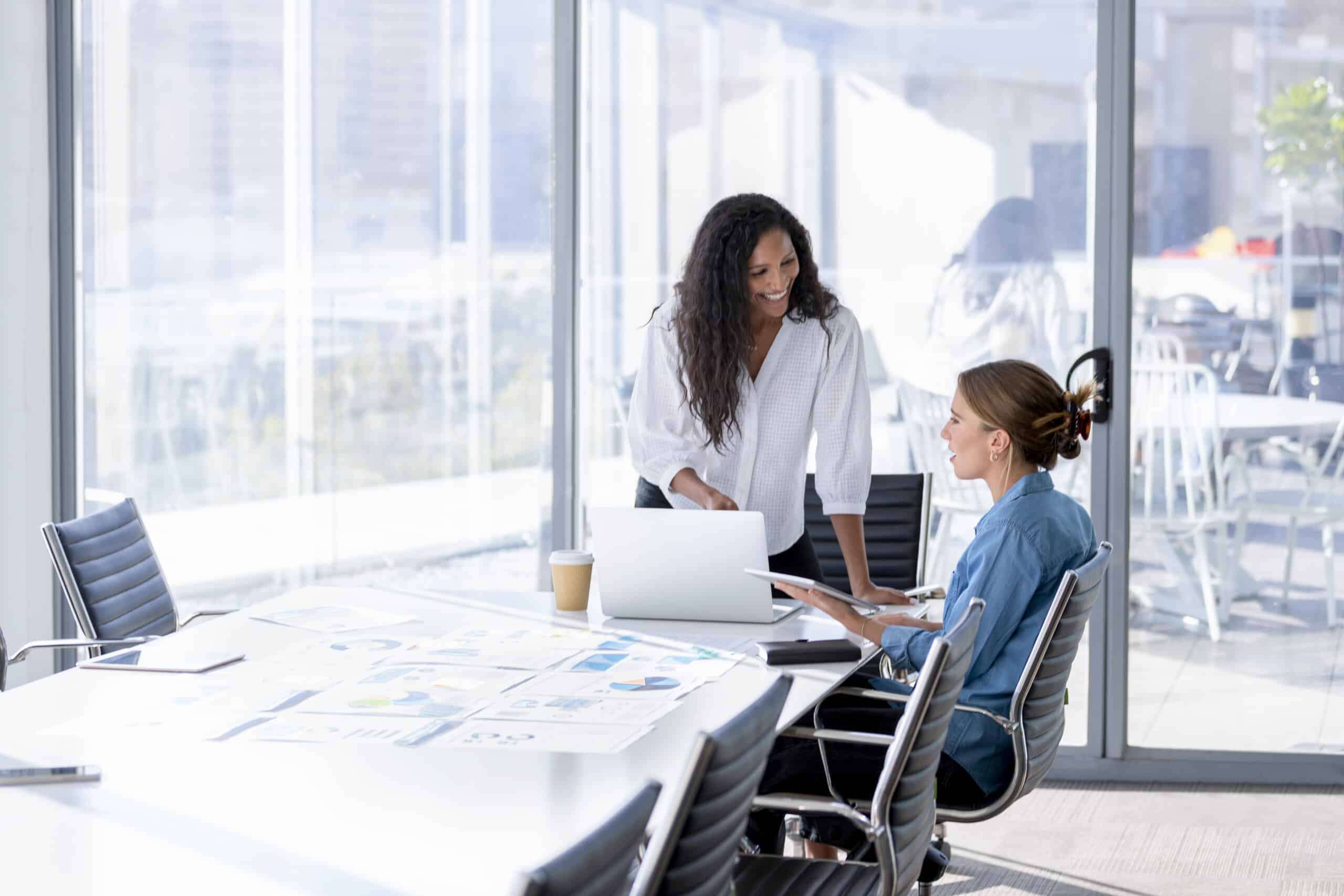 Two women discuss real estate fair market value analysis at a conference table in a bright office.